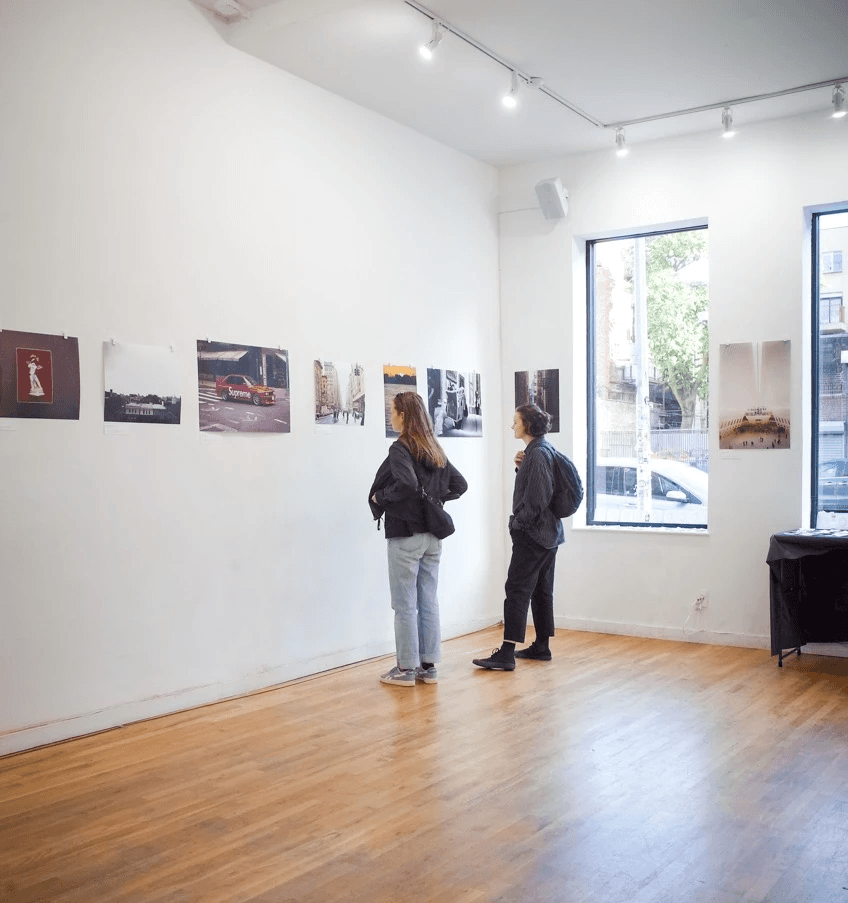 Two visitors viewing the “New York Narratives” prints in a minimal gallery with large front windows.