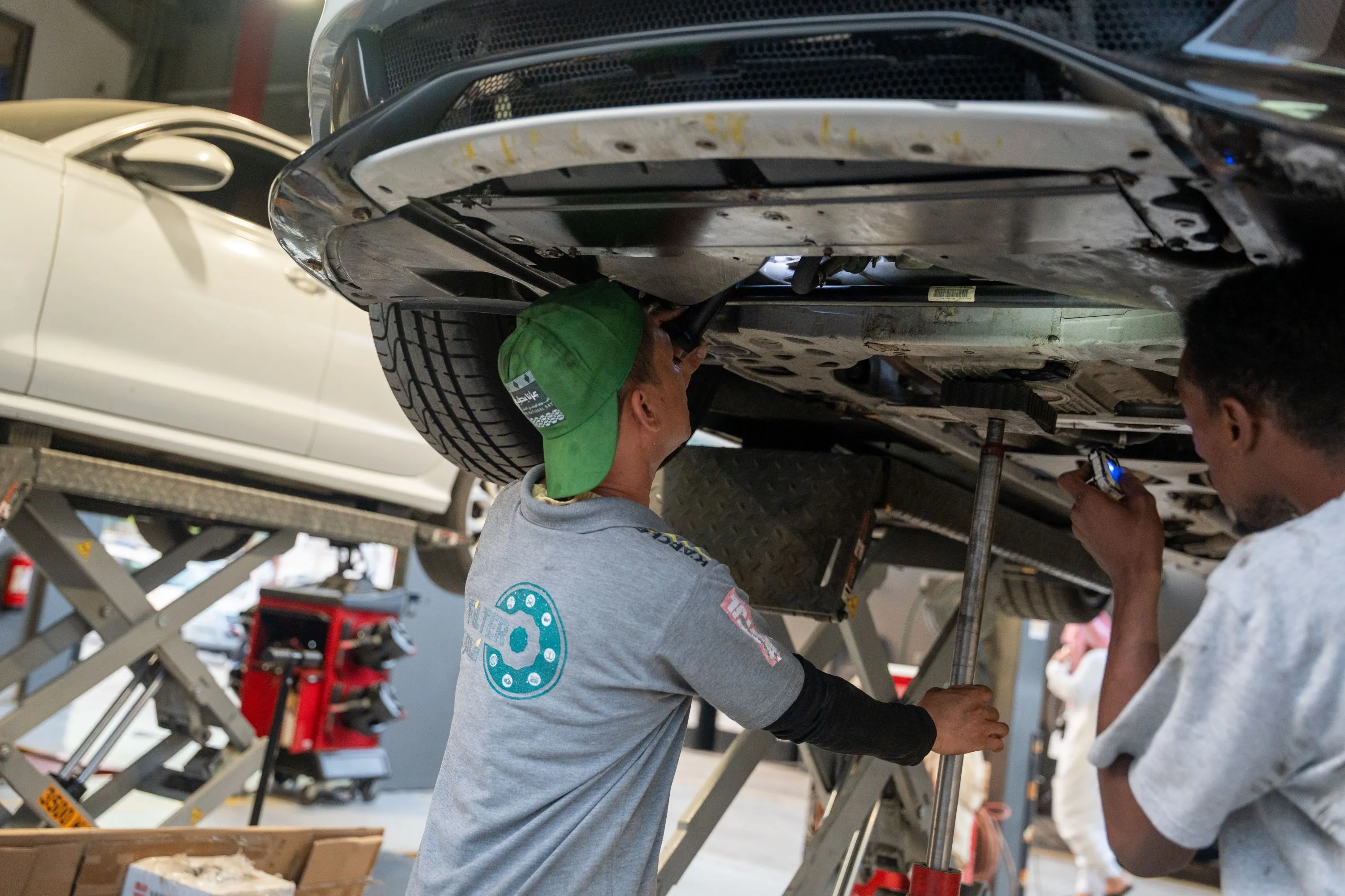 عامل يفحص أسفل السيارة مع دفتر الملاحظات في يده، ويرتدي معدات واقية وجاكيت.