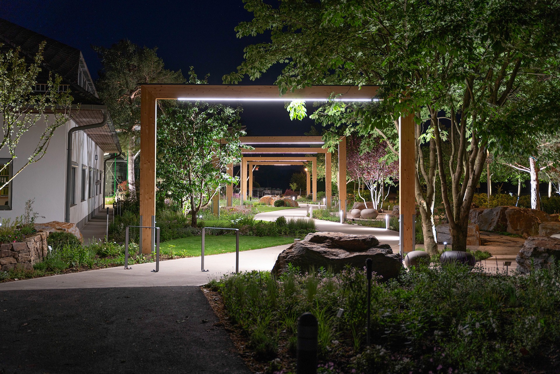 Beleuchteter Gartenweg mit Holzrahmen und üppigen Pflanzbeeten in einem Schaugarten bei Nacht.
