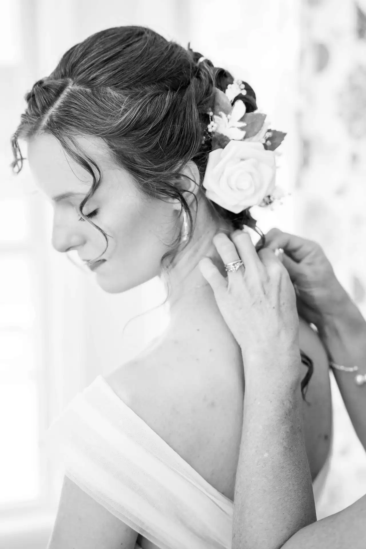 A black and white image of a woman with flowers in her hair and another woman gently putting her necklace together