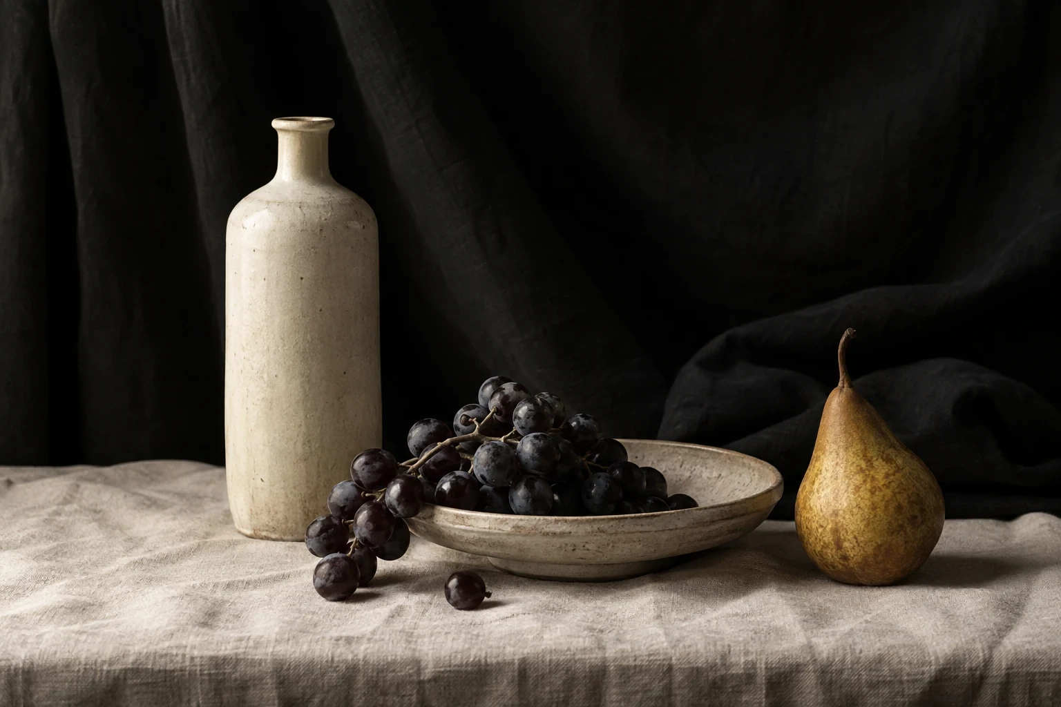 Ivory ceramic bottle, a shallow dish of dark grapes and a ripe pear on pale linen against black draped cloth