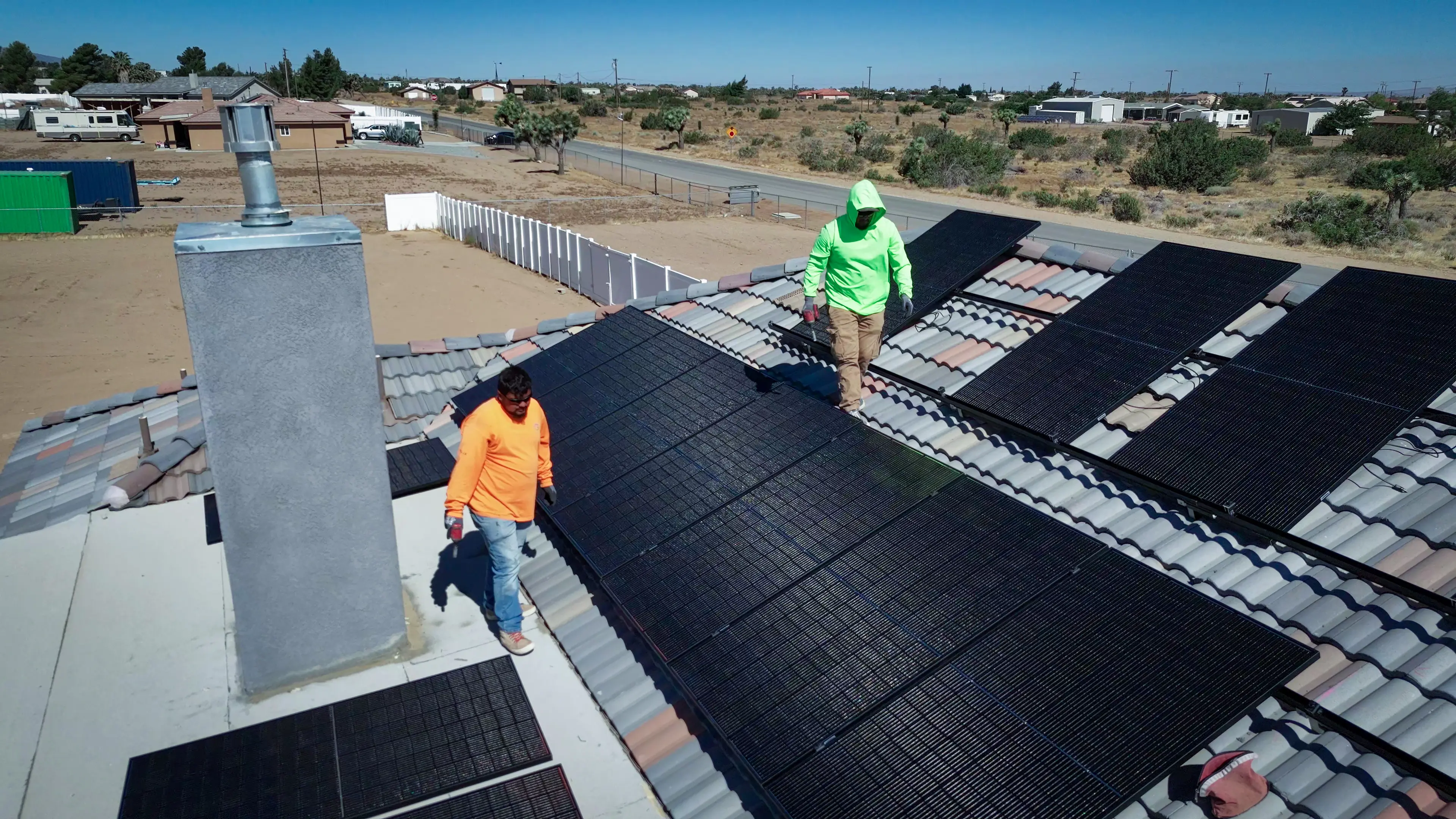 Two Option One Solar installers walking across a tile roof already covered with black solar panels, with High Desert scrub and houses behind them