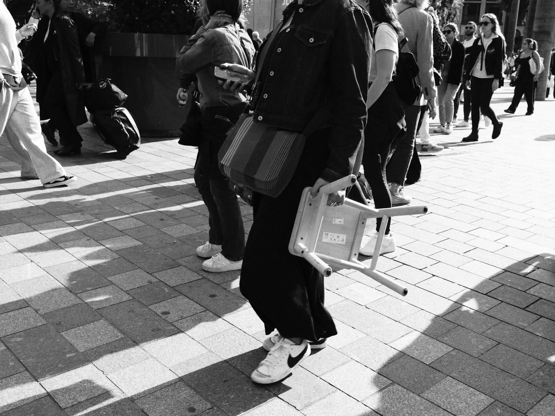 Person carrying a small chair through a crowd, in black and white