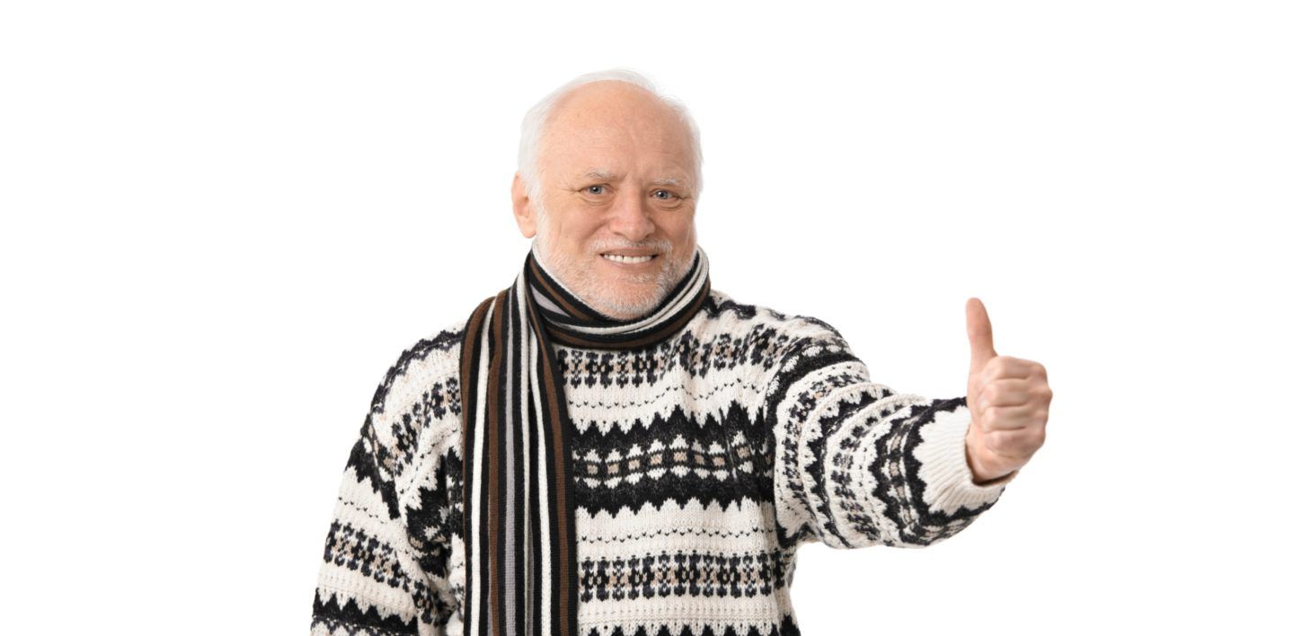 Uomo anziano con maglione di lana a motivi geometrici e sciarpa a righe posa su sfondo bianco, con il braccio teso in avanti e il pollice alzato in segno di approvazione.