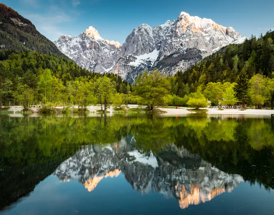 A perfect mirror reflection of the snow-dusted Julian Alps in the still, clear water of Lake Jasna, Slovenia, framed by vibrant green spring trees and a bright blue sky.