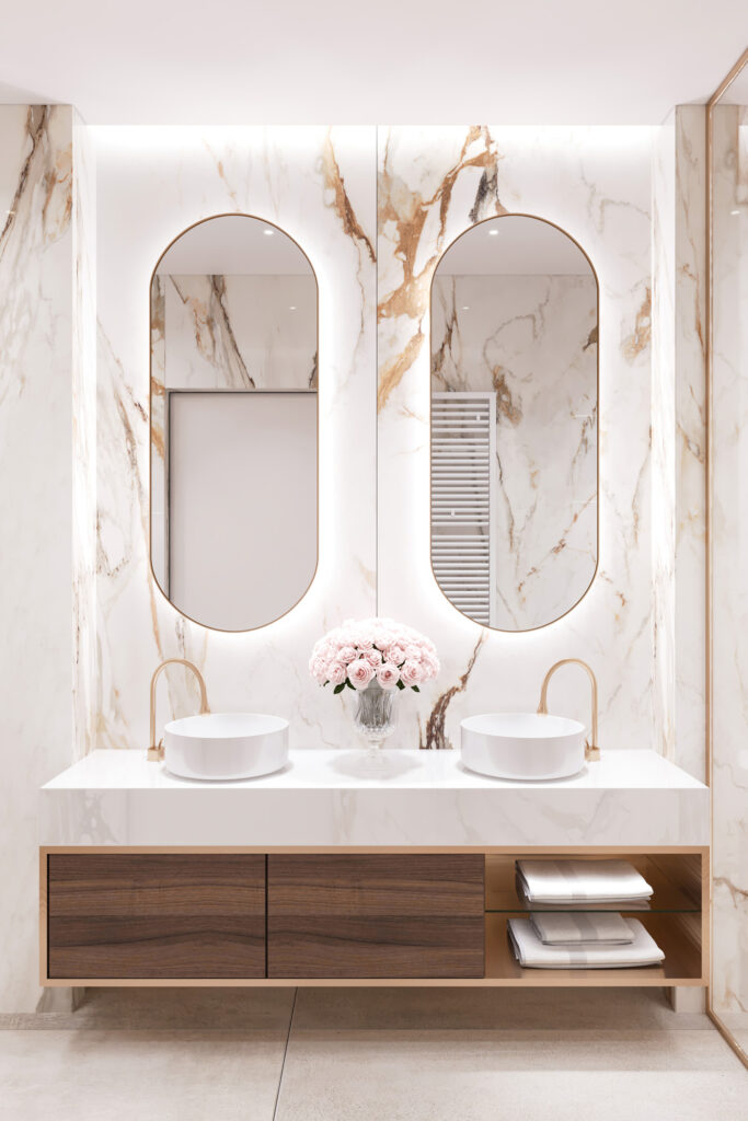 Modern bathroom with marble walls and countertops, two oval mirrors, dual white vessel sinks with gold faucets, and a vase of pink roses centered between the sinks