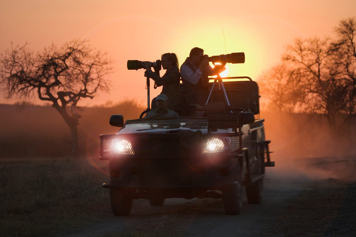 Photographers with long lenses on an open vehicle, silhouetted against a low red sun