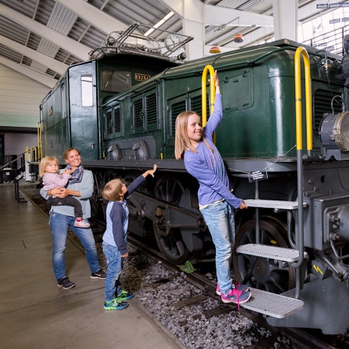 A woman with three children explore an old green train in a museum. One child climbs steps, and another points at the train.
