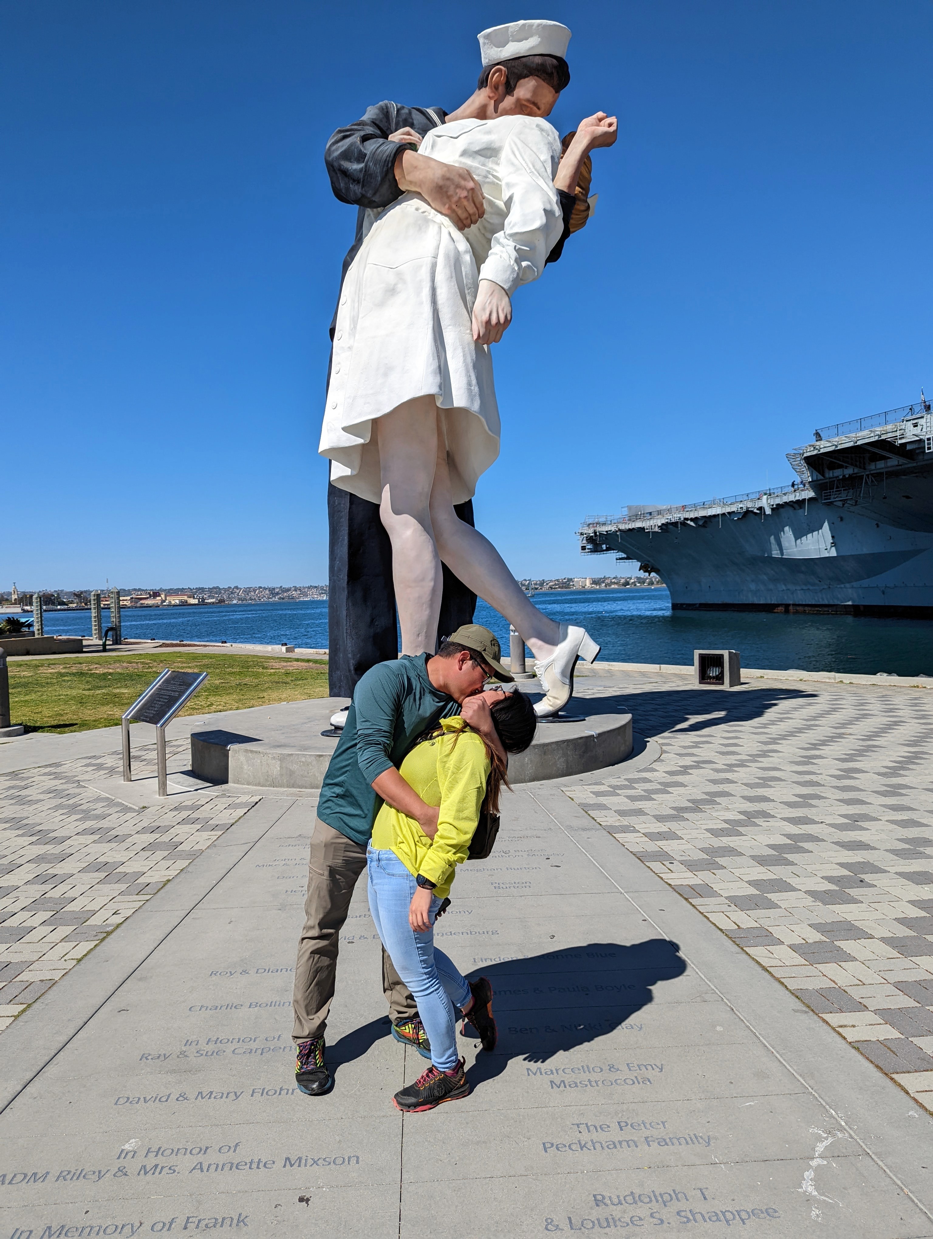 A couple holds hands, standing on a rocky beach under a large cave arch, gazing at the tranquil ocean during sunset. The sky is softly lit in pastel hues.