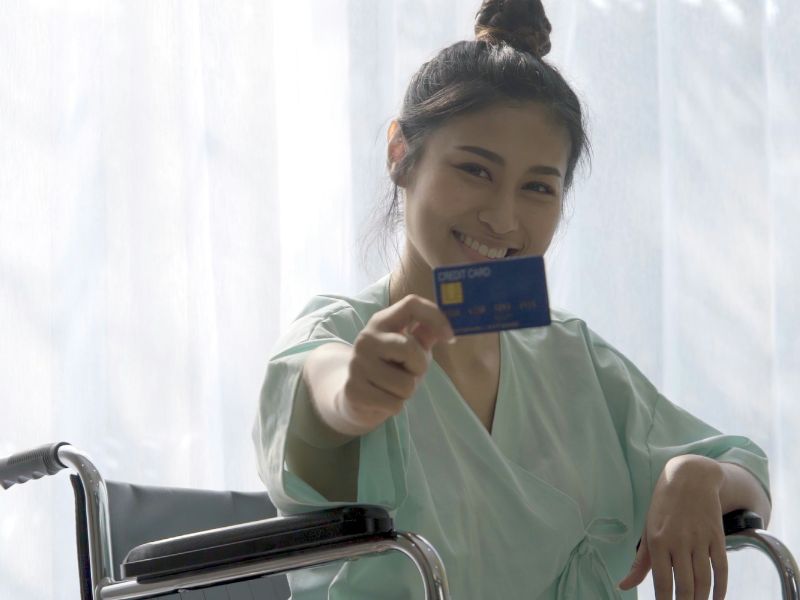 A smiling woman in a wheelchair, offering a credit card to the camera, highlighting financial security and support.