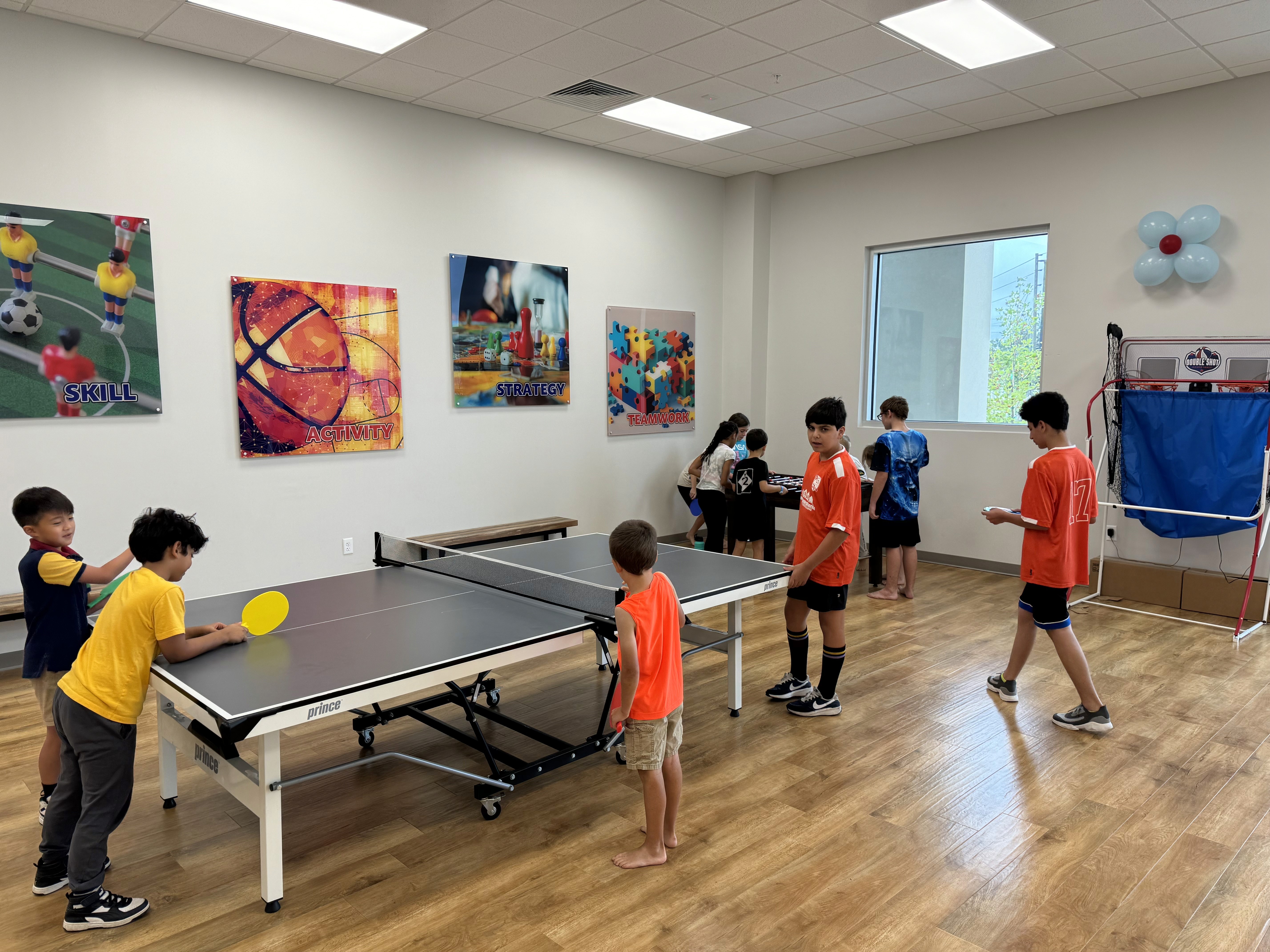 A group of kids playing table ping pong and hanging out together in a bright activity room.