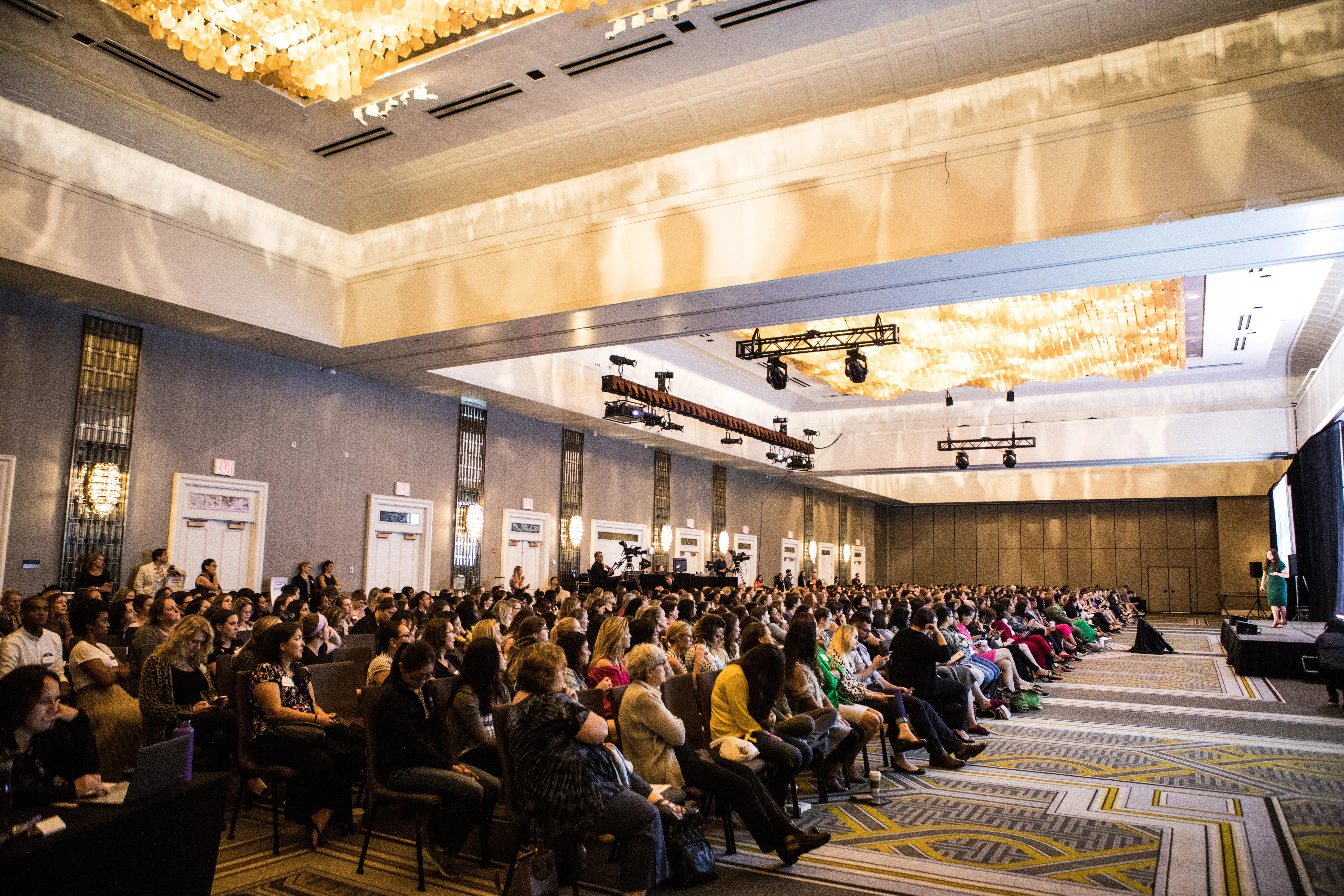 A large gathering of people is seated in rows in a spacious, elegantly decorated conference hall with chandeliers and a patterned carpet, attentively watching a presentation on stage.