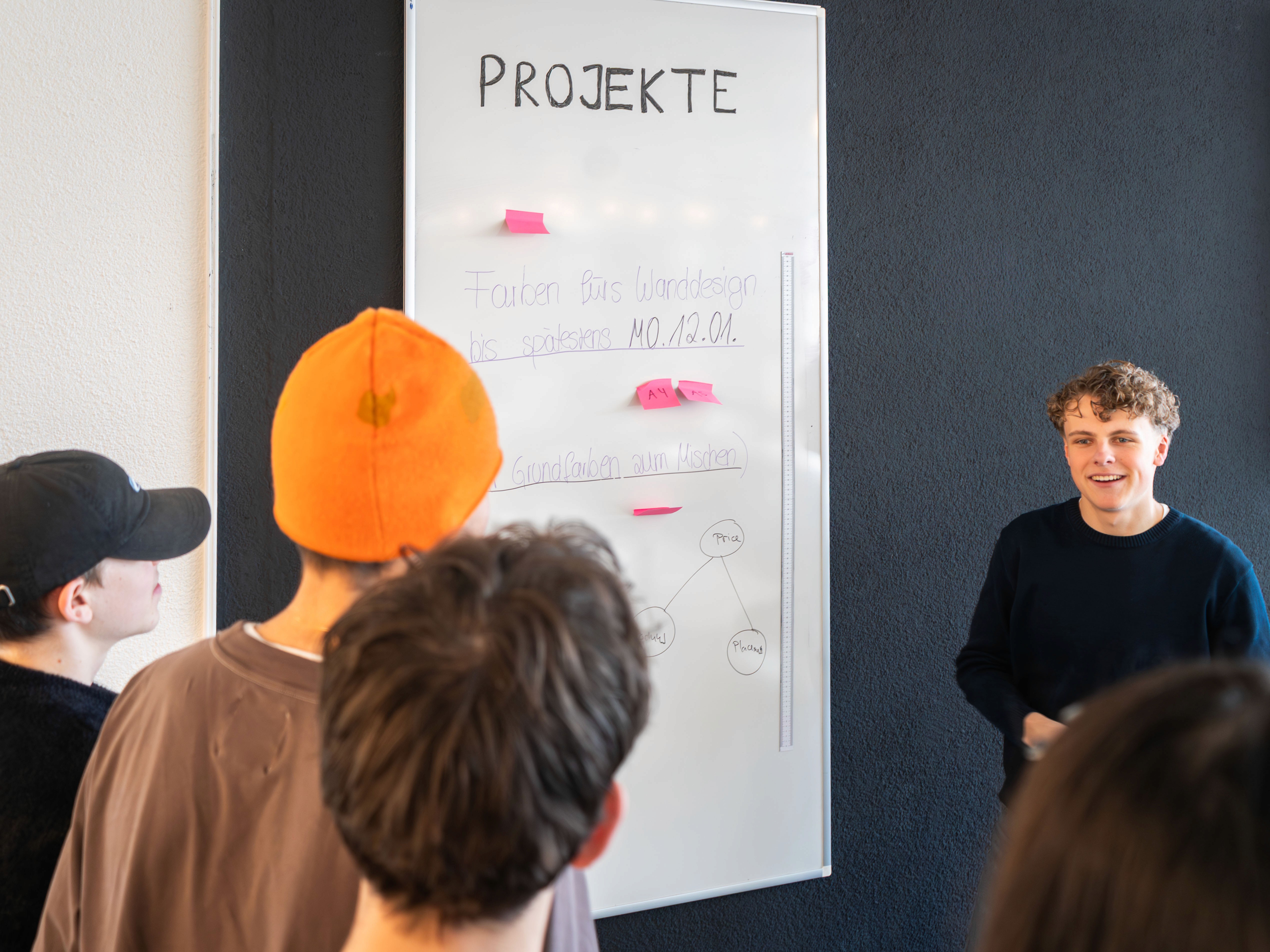 A young person with curly hair smiles at the camera while sitting in front of a laptop against a dark background.