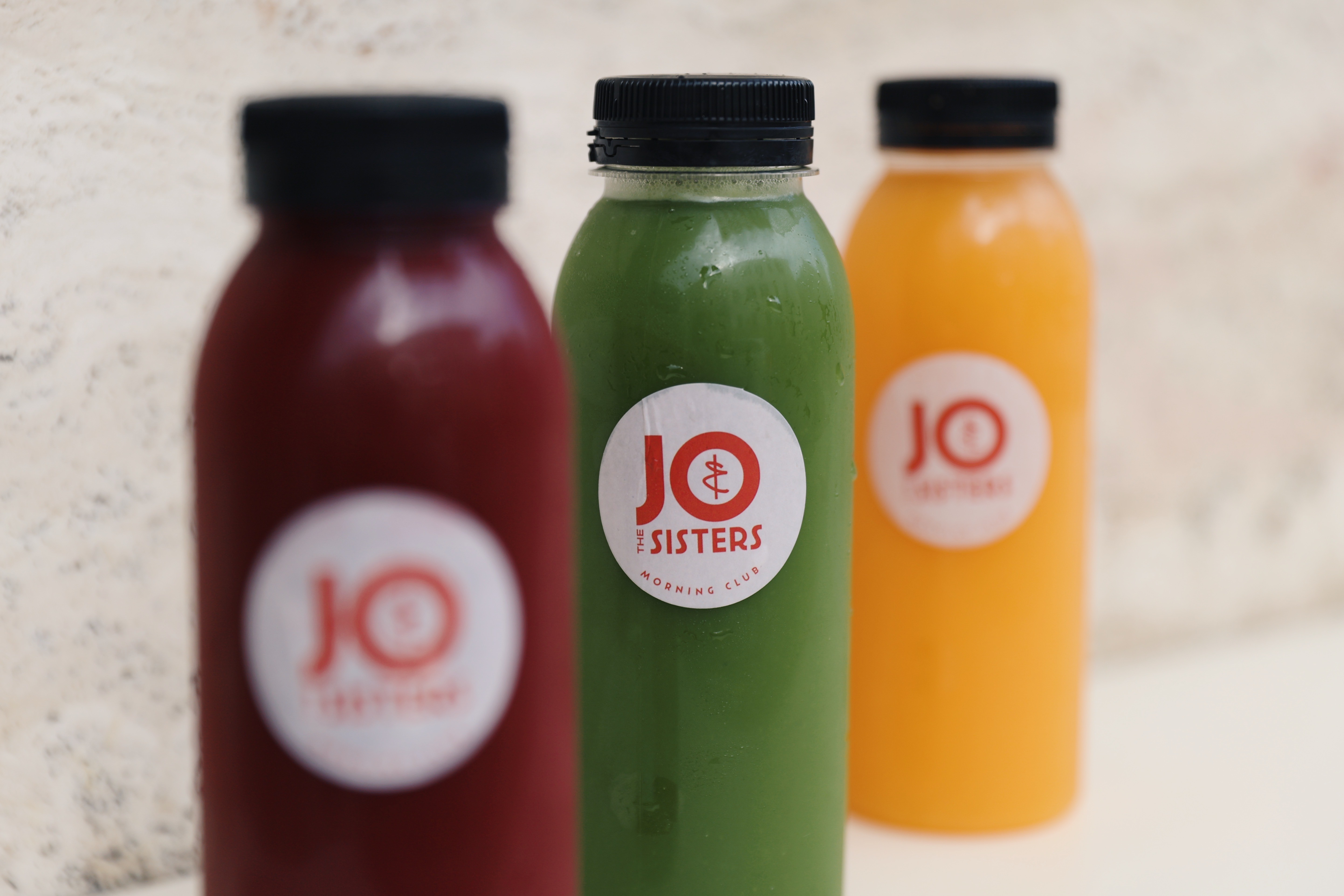 Three cold-pressed juice bottles from Jo & The Sisters in red, green, and orange, arranged in a row with the green bottle in focus.