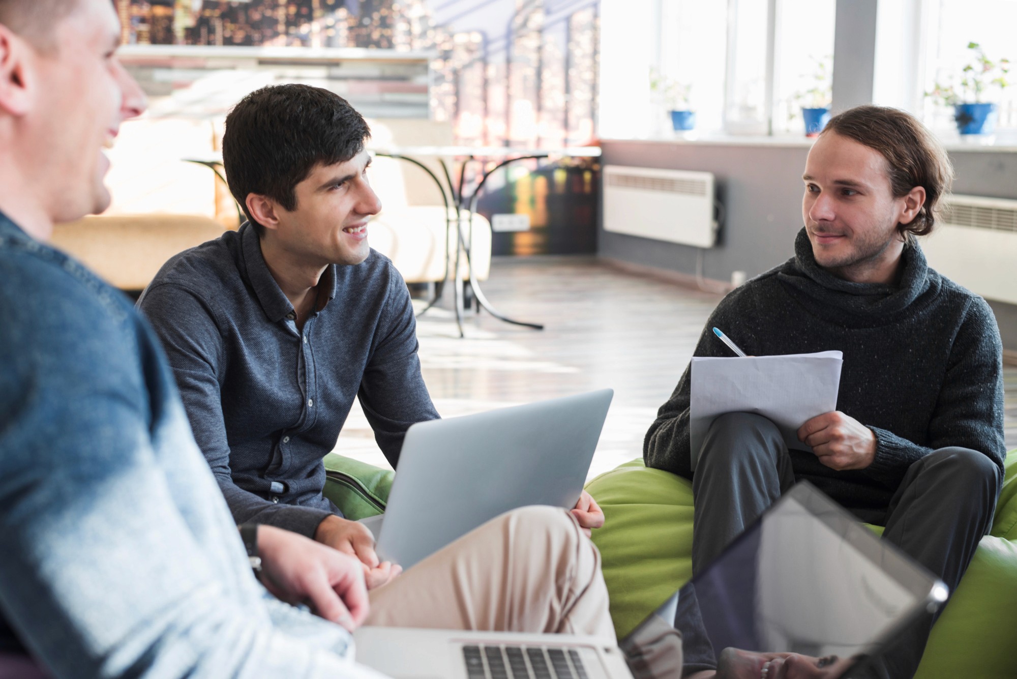 Small group discussion in a casual office lounge, colleagues talking with laptops and notes