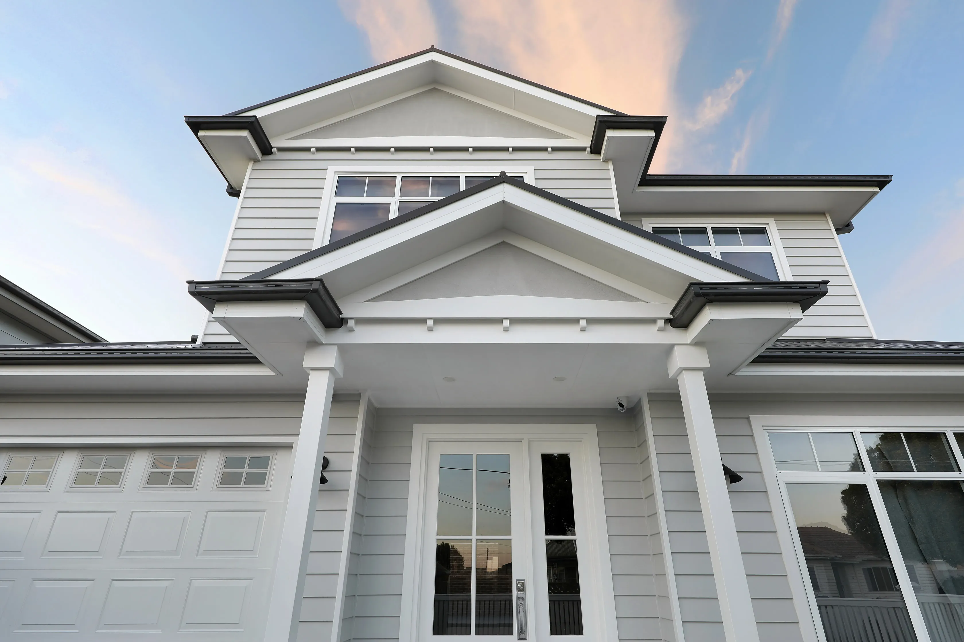 Upward view of the Lalor Residence entry, showing the stacked white gabled portico, weatherboard cladding, dark gutters and a glazed white front door at dusk