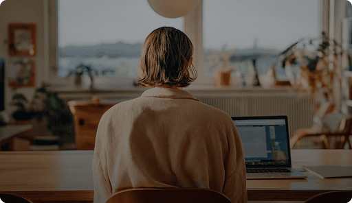 A person with short hair is sitting at a desk in a softly lit room, working on a laptop.