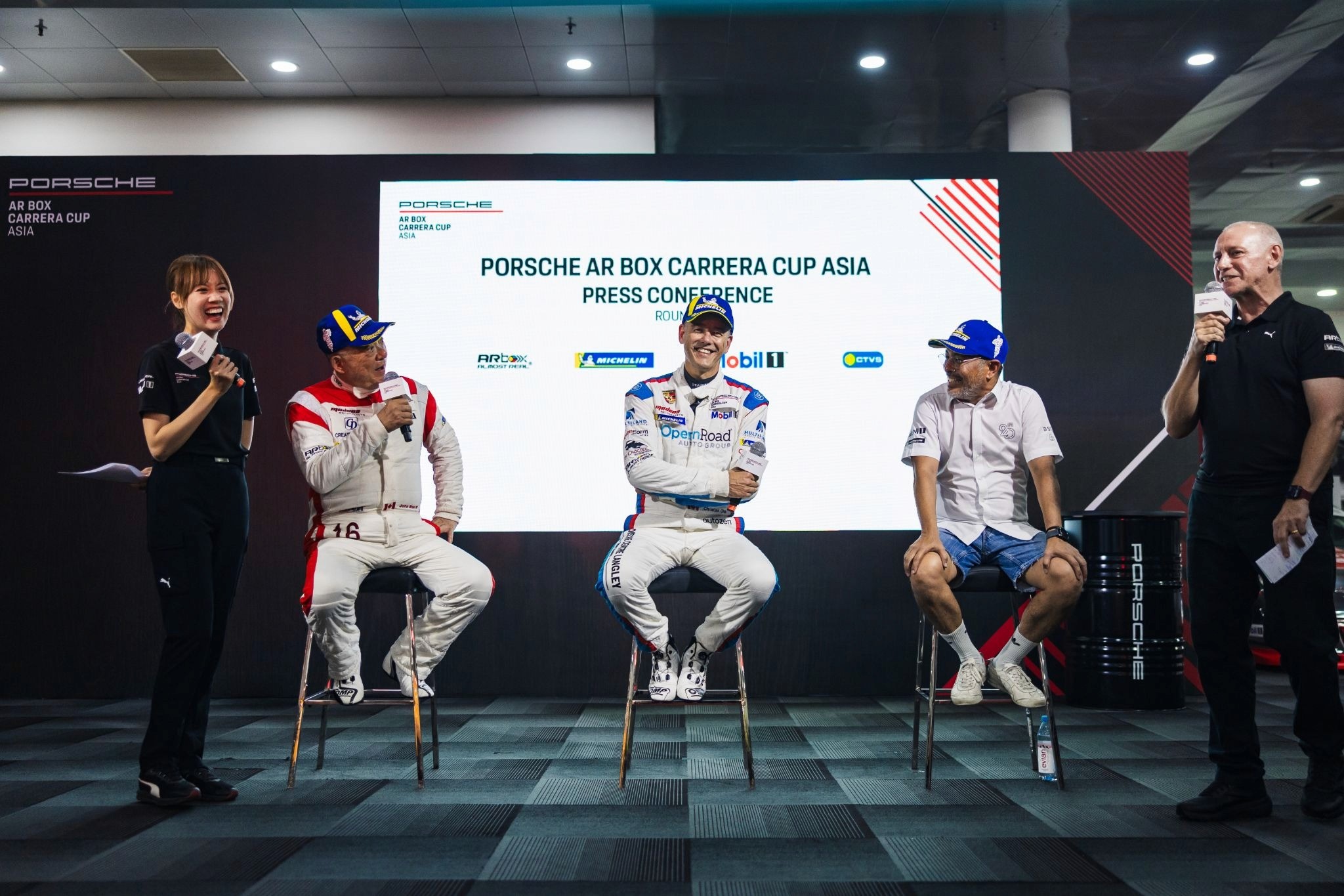 Presenter Sammie Hum stands with a microphone on a stage during a Porsche AR Box Carrera Cup Asia press conference, laughing alongside seated racing drivers and co-host Steve Dawson in front of a white media backdrop.