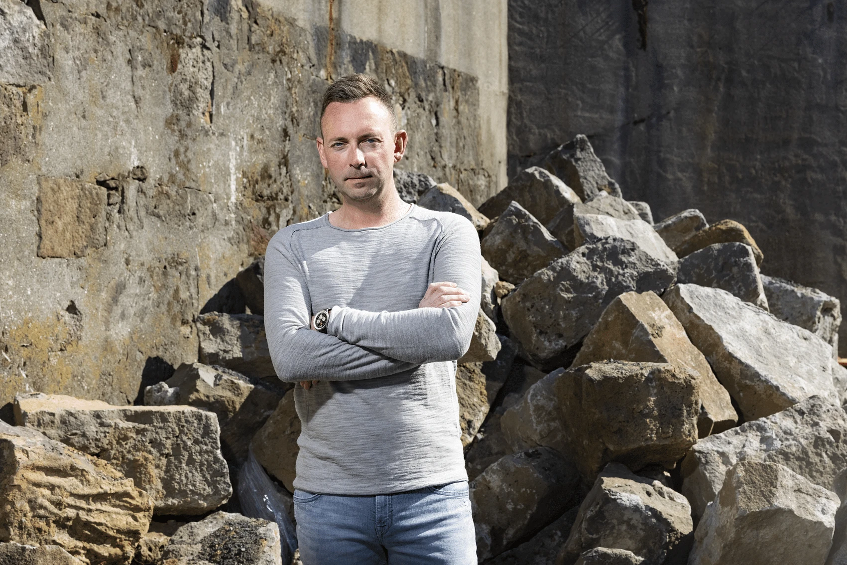 Author Ragnar Jónasson, co-founder of Iceland Noir, looks to one side with his chin resting on his hand, seated outdoors in front of weathered stone steps and a wooden wall.