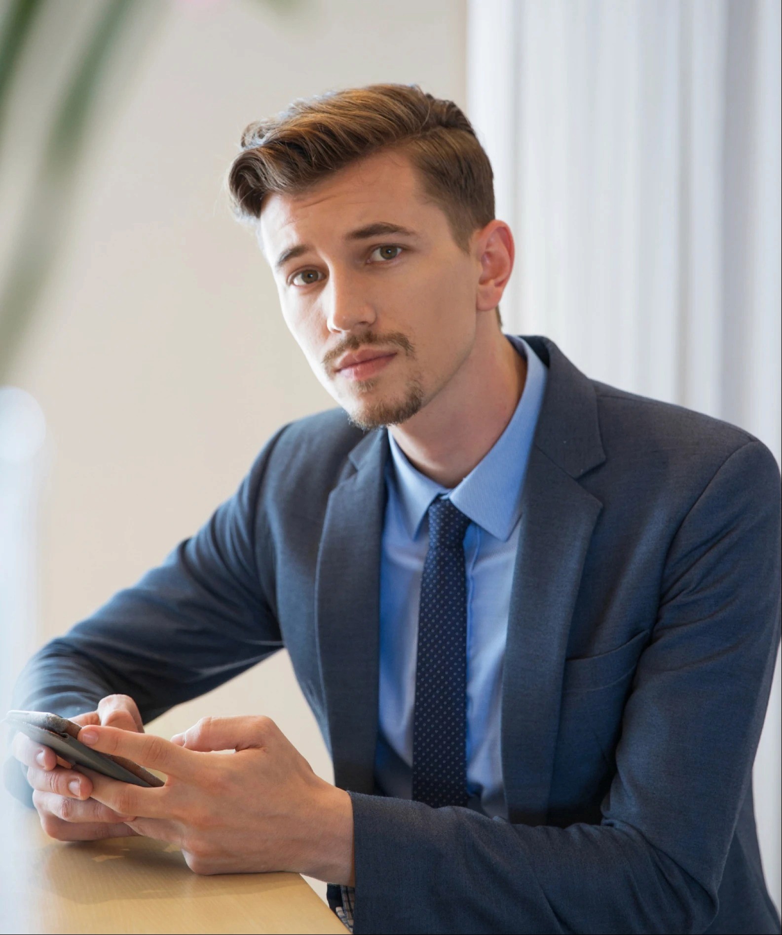 A man in a suit sits at a table, looking thoughtfully at his smartphone, with natural light in the background.