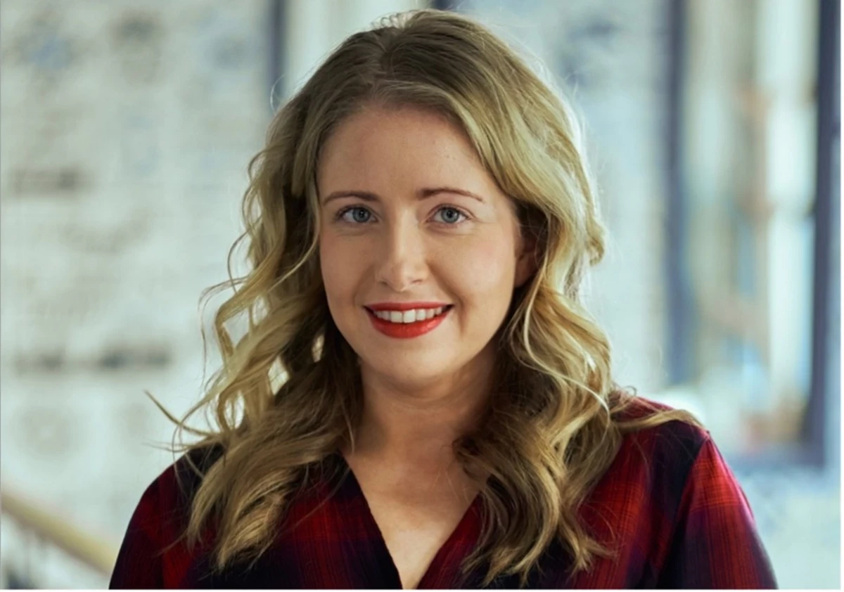 A person with long, wavy blonde hair stands indoors, wearing a red plaid shirt against a softly blurred background.
