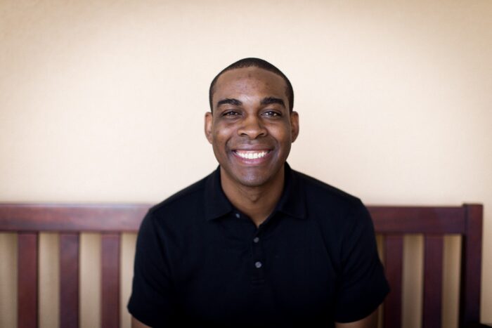 Smiling man in a black polo shirt sits on a wooden bench against a plain beige wall, facing the camera in a relaxed, friendly pose.