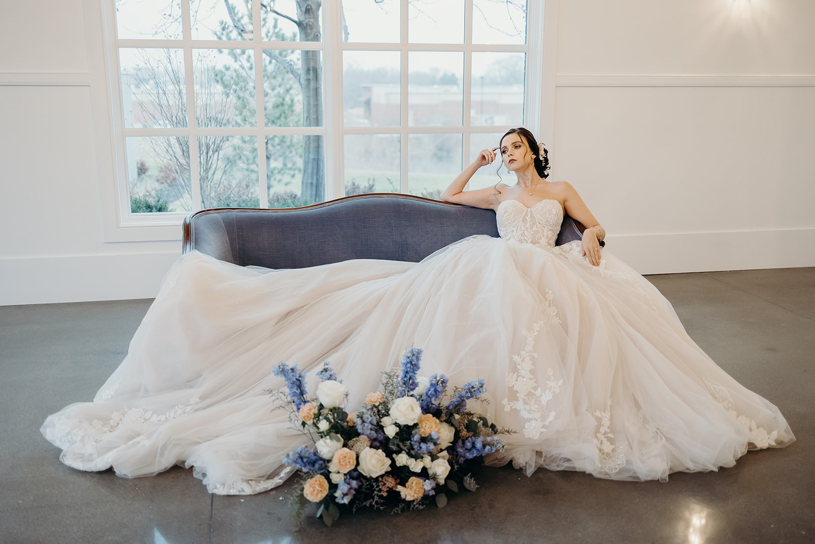 Bride in strapless ball gown wedding dress with lace details, sitting on blue velvet sofa with pastel floral bouquet.