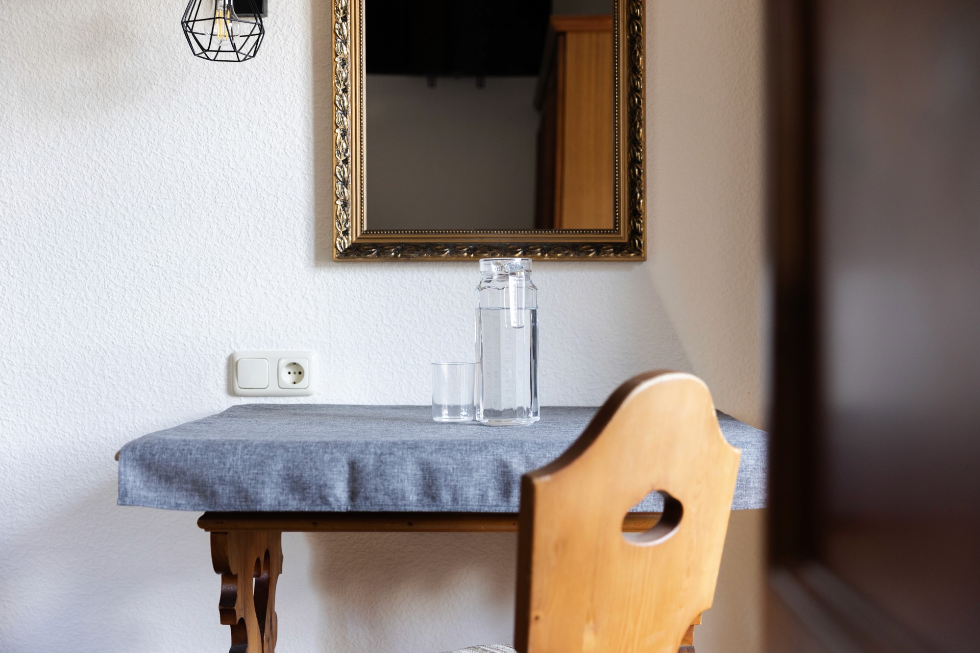 Auf einem kleinen Holztisch mit grauer Tischdecke stehen eine Flasche aus Klarglas und ein Glas, platziert unter einem dekorativen Wandspiegel in einem gemütlichen, gut beleuchteten Raum.
