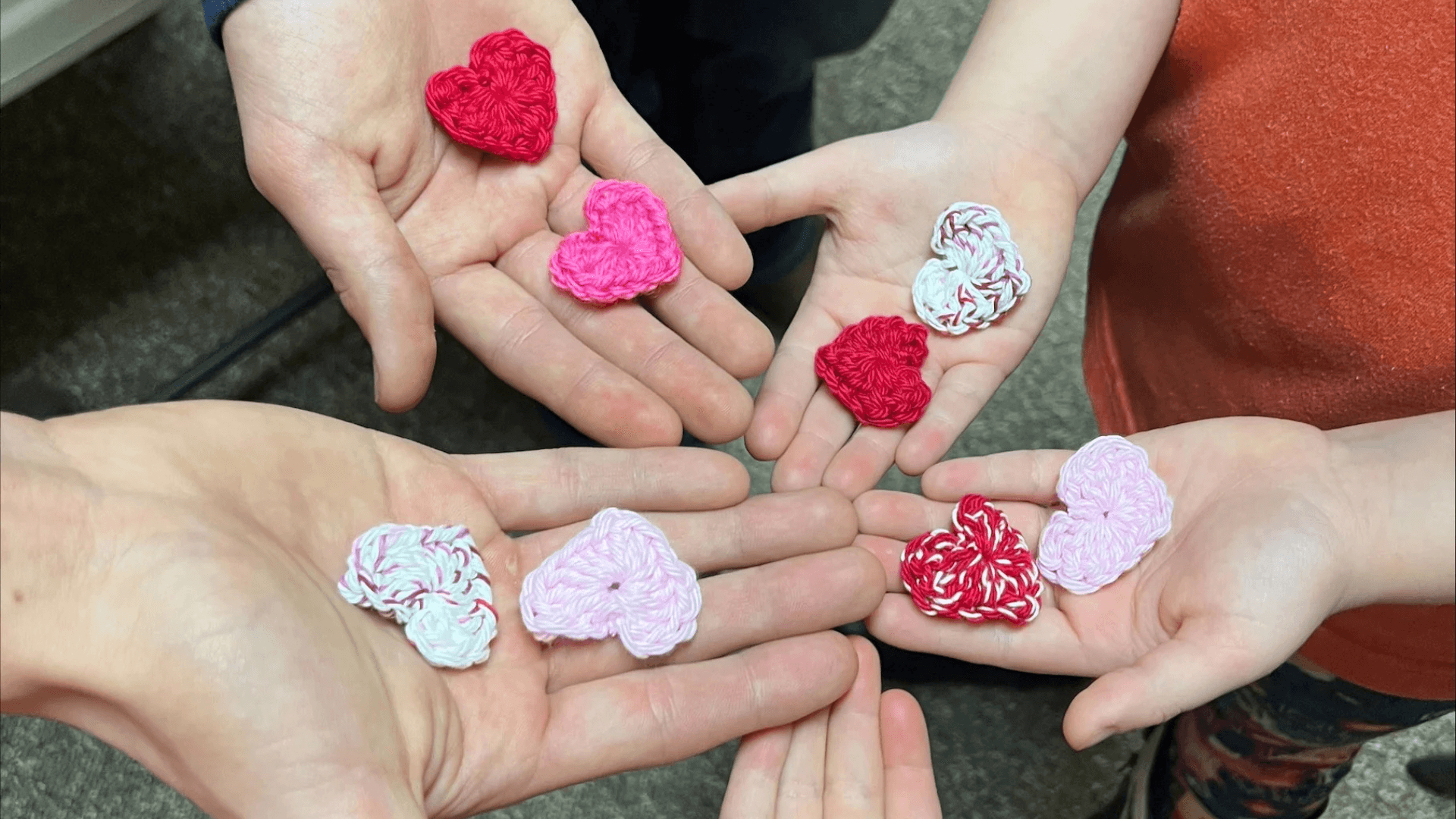 Alt tag: Close-up of several hands holding small crocheted heart shapes in red, pink, and white.