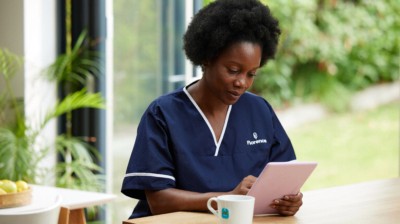 Healthcare worker in Florence scrubs using a tablet at a table with a cup of coffee in a bright indoor setting.