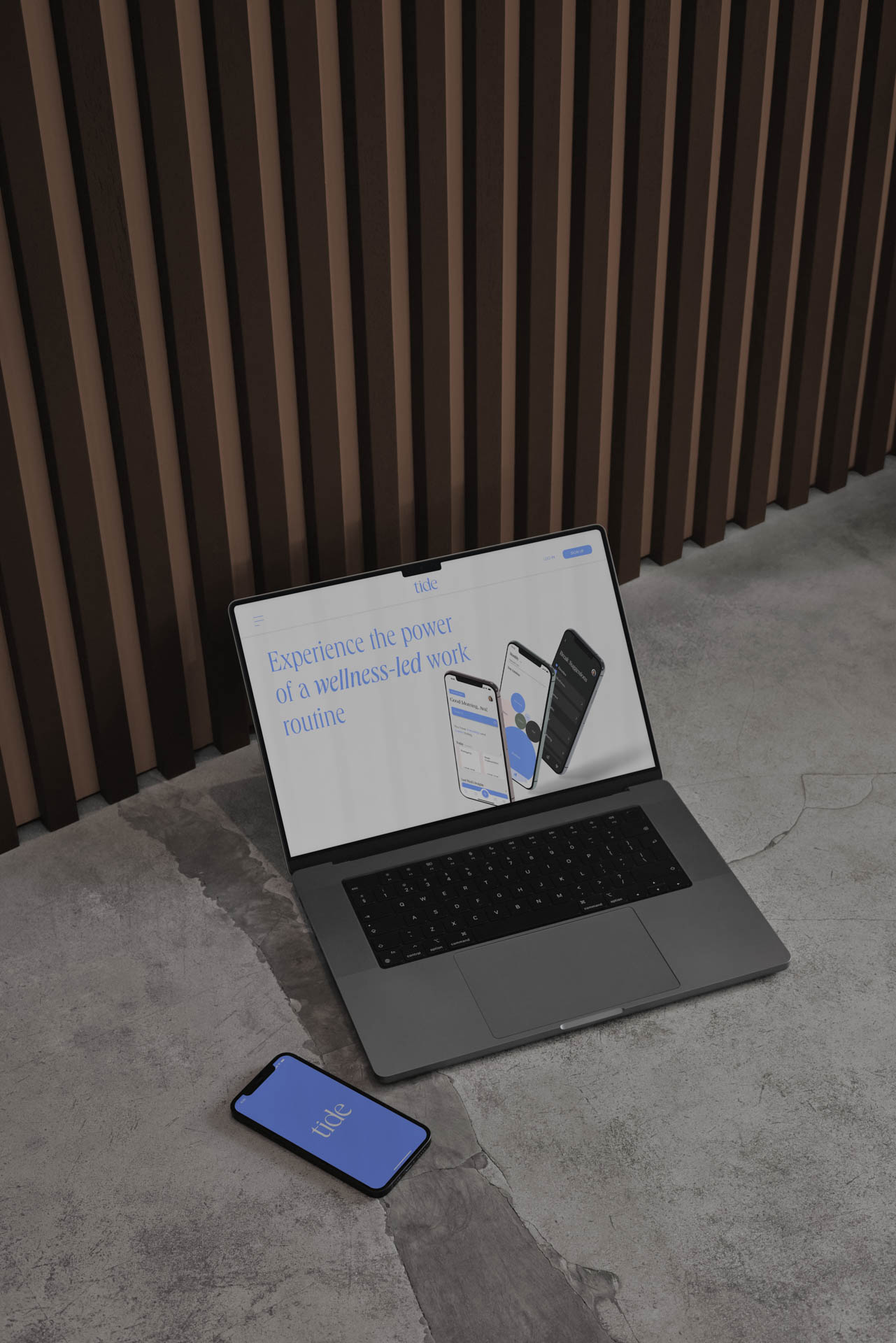 A dark gray laptop is open on a gray concrete surface, displaying a webpage titled “Experience the power of wellness-led work routine.” Three smartphones are shown on the laptop’s screen: one white, one light blue, and one dark gray. The laptop is positioned near a black smartphone with a blue screen displaying “Lite,” and a black smartphone with a white screen displaying “Lite” is partially visible behind the laptop. A vertical striped wall in dark brown and beige tones forms the backdrop.