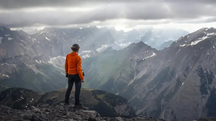 Personne en veste orange, debout sur un sommet rocheux, contemplant un vaste paysage de montagnes brumeuses et vallées. Ambiance d’aventure et de réflexion en altitude, luminosité sombre et atmosphérique