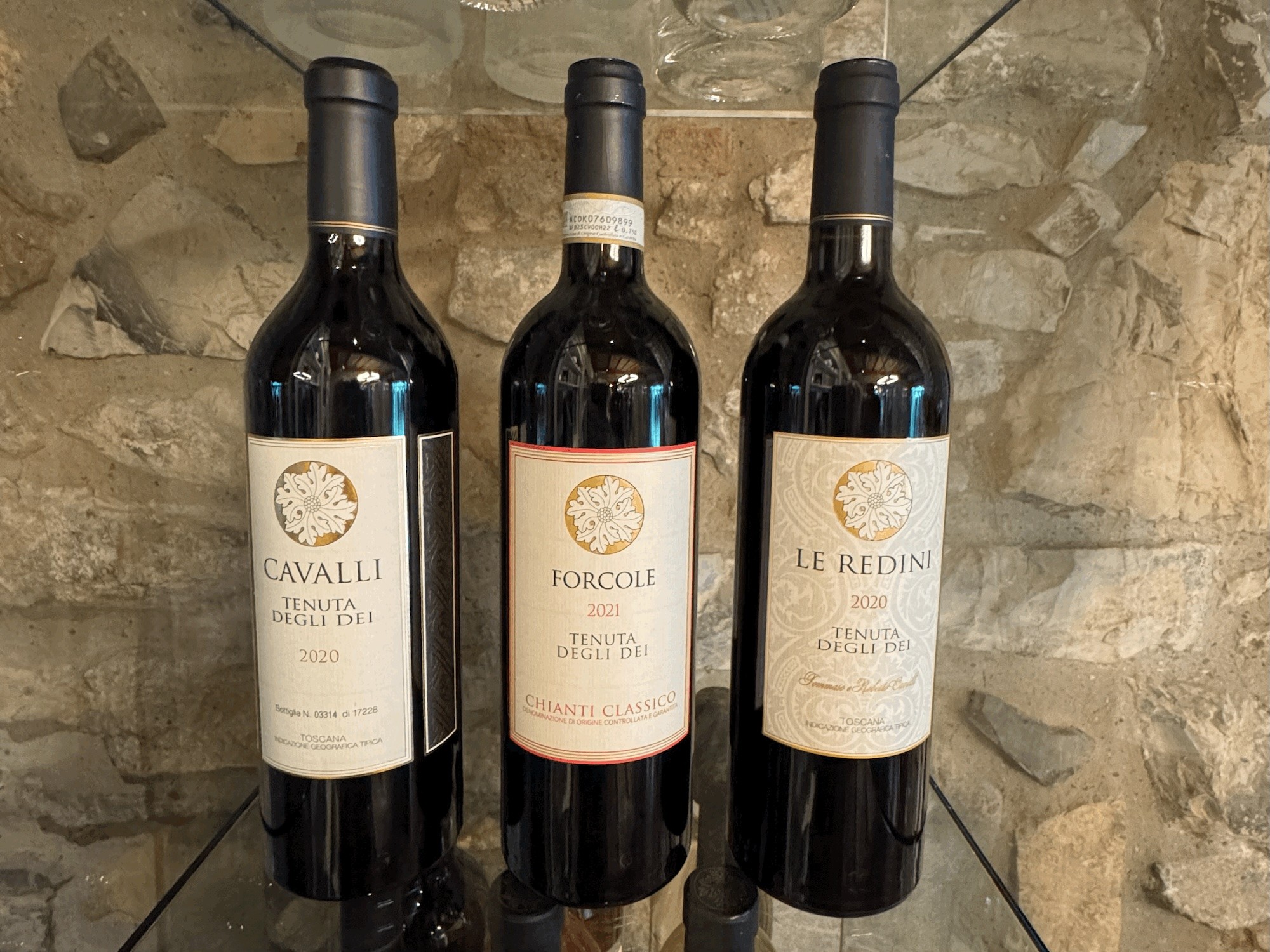 Bottles of 2020 Cavalli, 2021 Forcole, and 2020 Le Redini displayed against a stone wall at Tenuta Degli Dei.