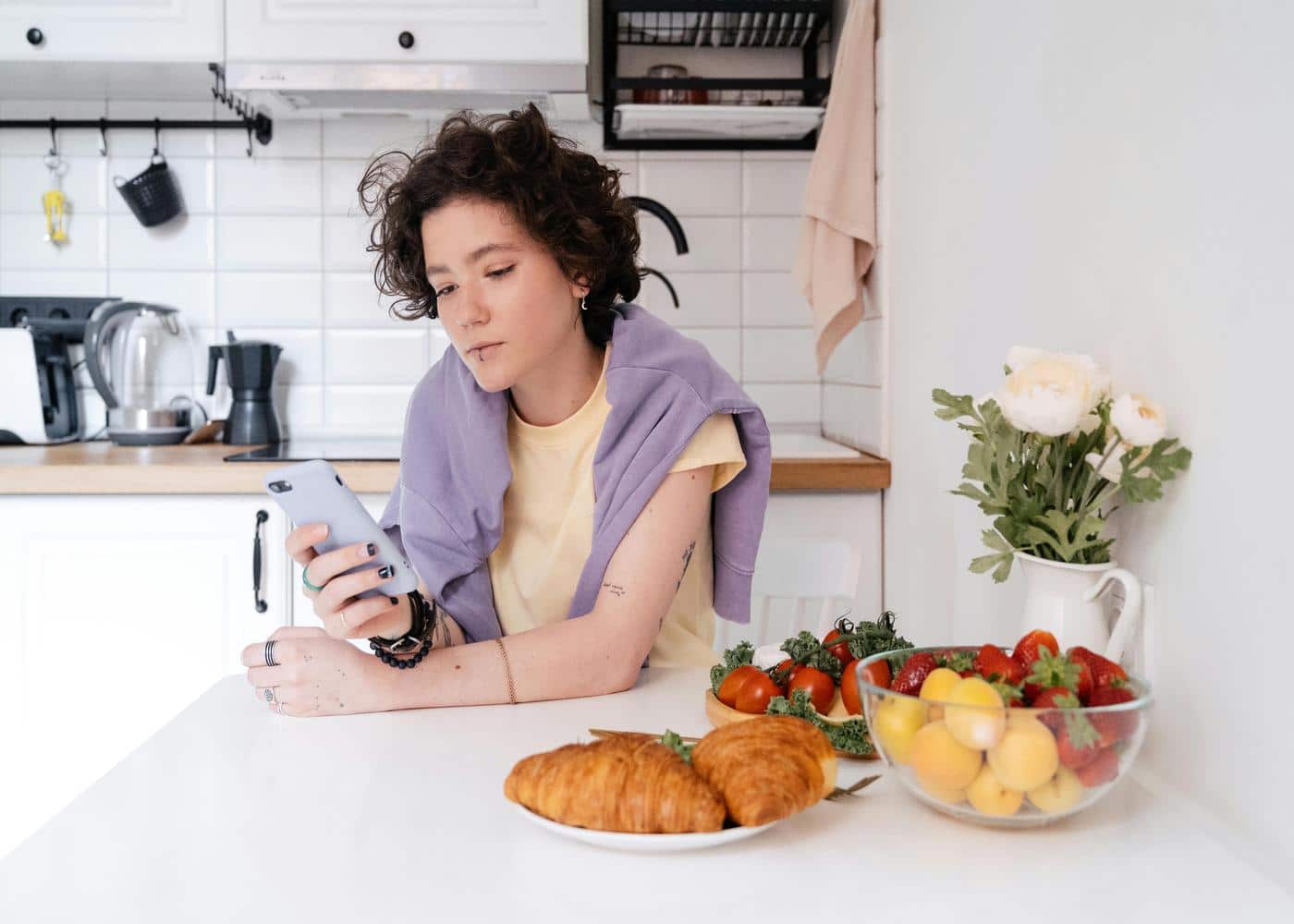 Woman sitting in kitchen with croissants and fruit next to her staring at smartphone