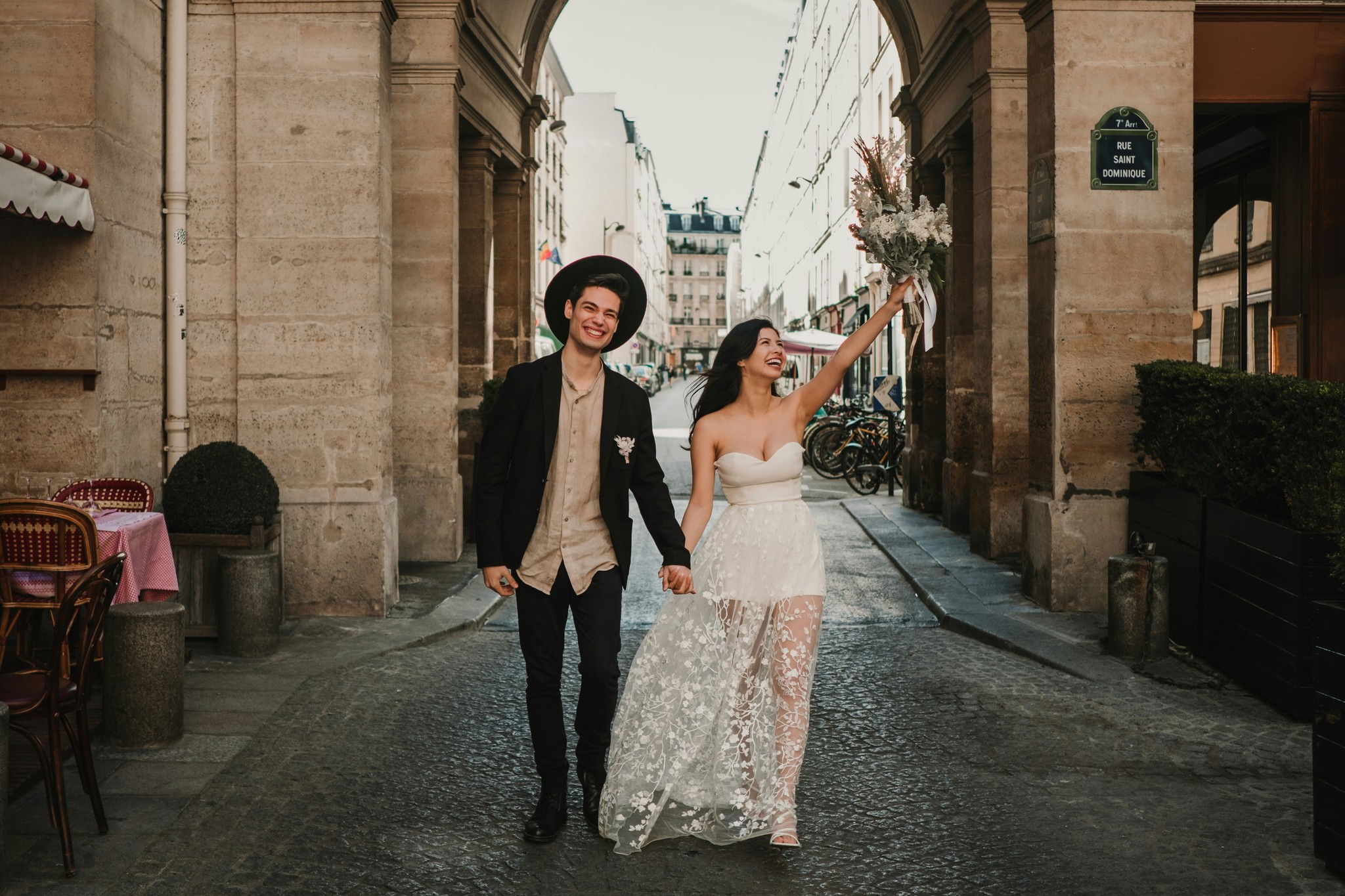 Laughing couple walking through a stone archway on a Parisian cobblestone street with the bride raising her dried flower bouquet