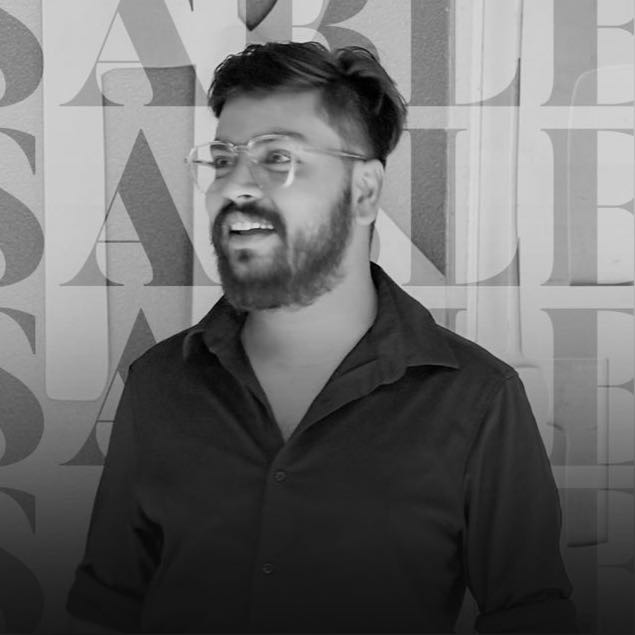 Person wearing a blue puffer jacket and red backpack standing in front of a light blue building on a city street.