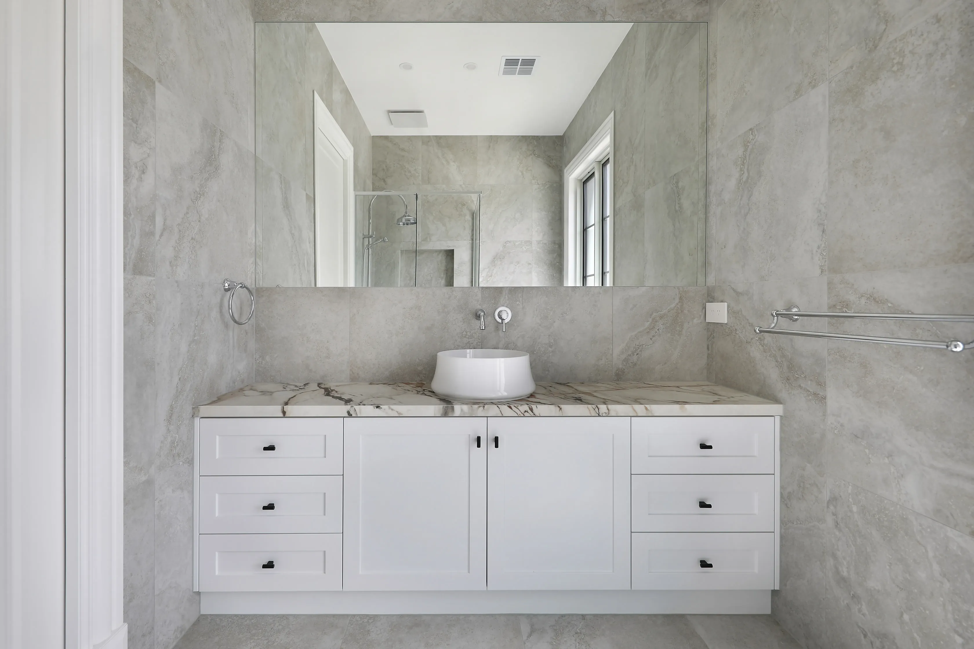 Bathroom in grey stone-look tiles with a white shaker vanity, marble benchtop, white vessel basin, large frameless mirror and chrome fittings
