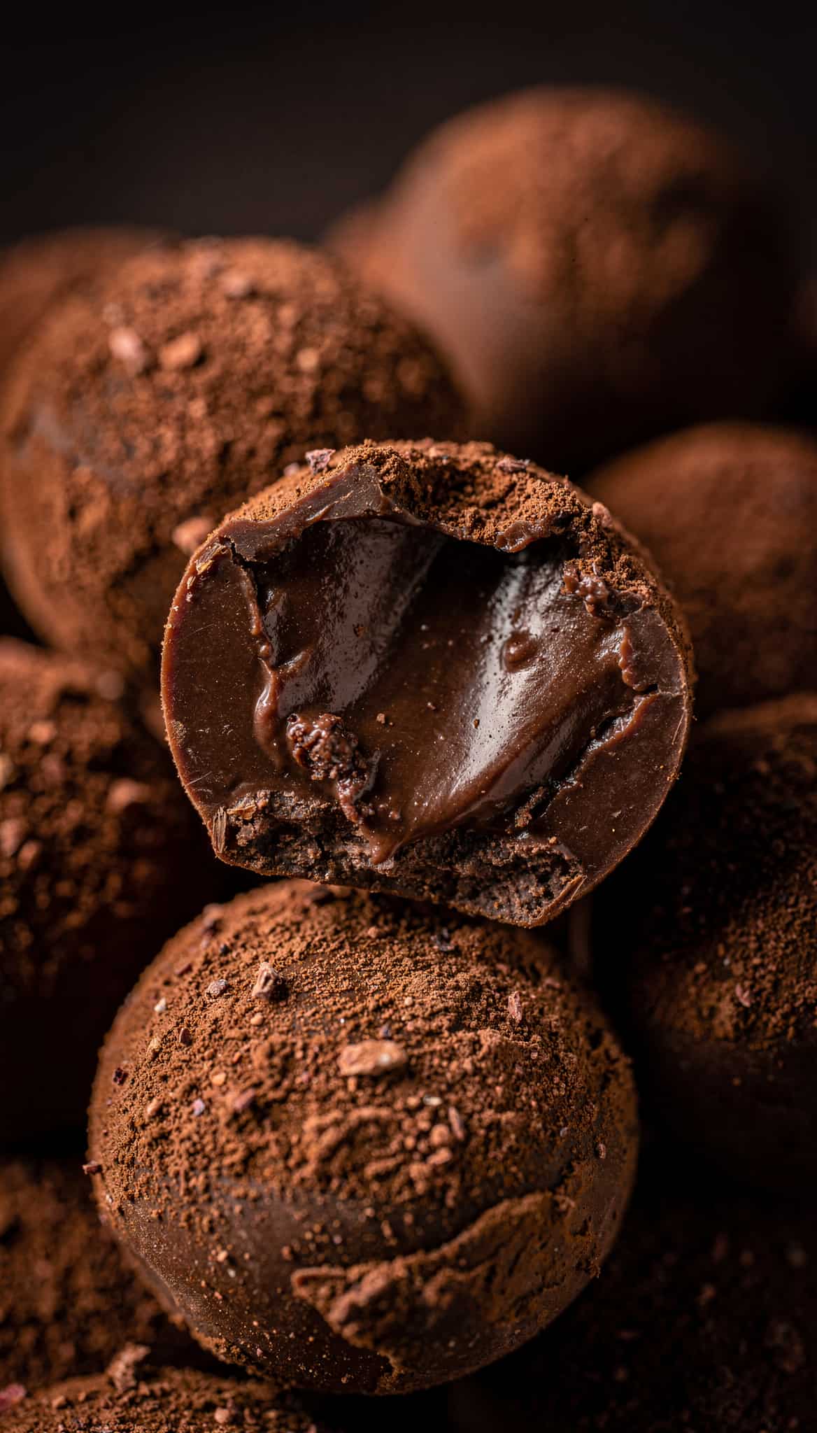 a pile of chocolate truffles dusted with cocoa powder. One truffle in the foreground is broken in half, revealing a rich, molten chocolate interior