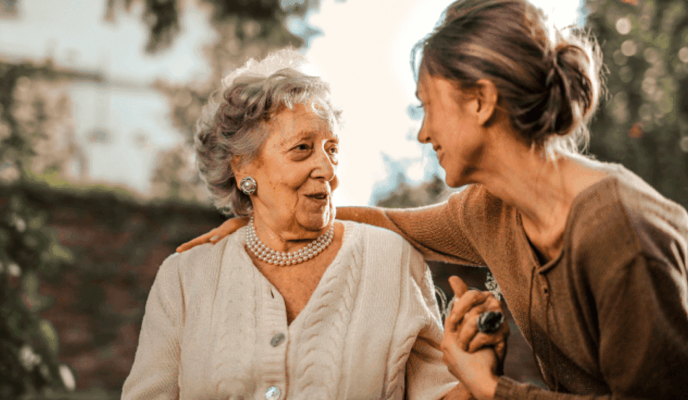 A younger woman leans in warmly toward an elderly woman wearing a pearl necklace and white cardigan, gently holding her hand and placing an arm around her shoulder in an outdoor setting.