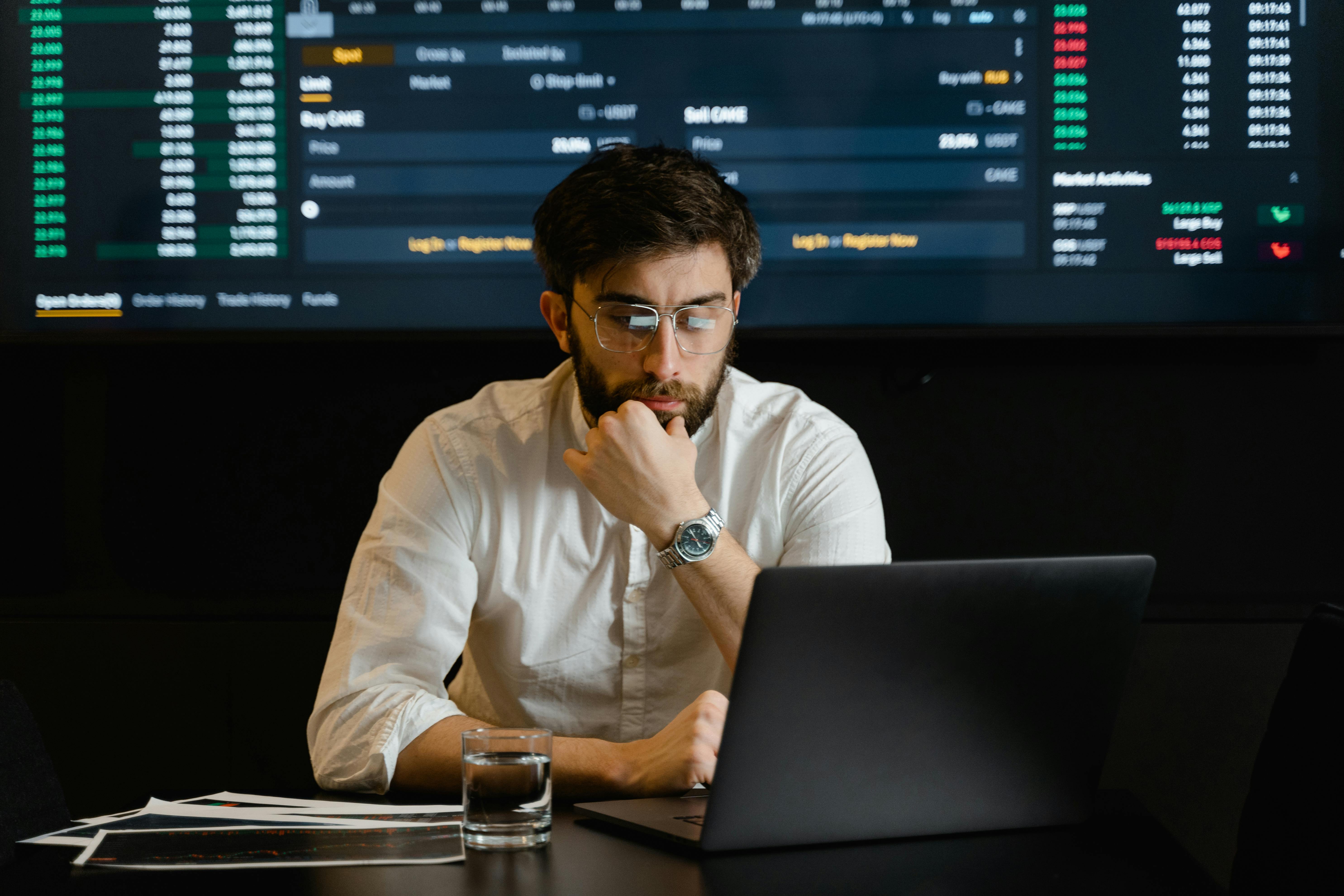 A man wearing glasses and a brown cardigan focuses intently on a laptop screen in a dimly lit room, conveying concentration and a pensive mood.
