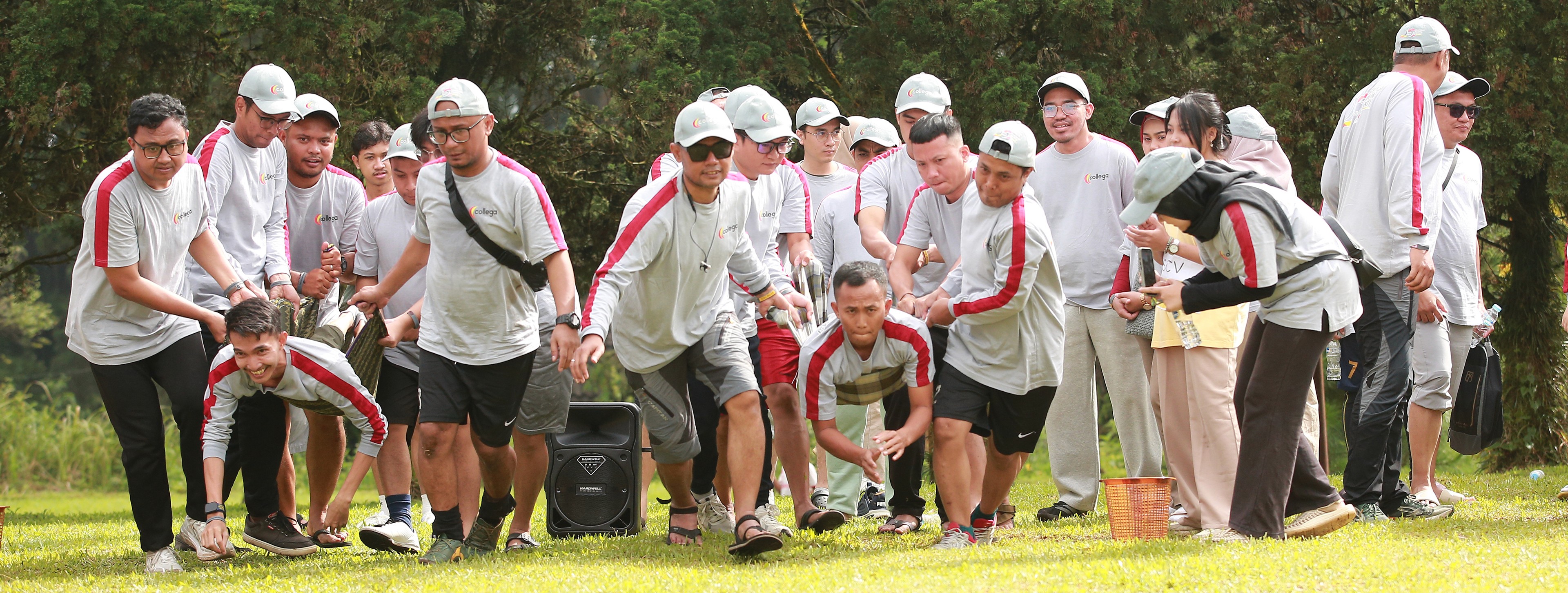 Beberapa peserta mengenakan kaos hitam dan oranye duduk berbaris di rumput sambil menuangkan tepung ke kepala rekan di belakangnya sebagai bagian dari permainan estafet, dengan ekspresi ceria dan suasana penuh tawa