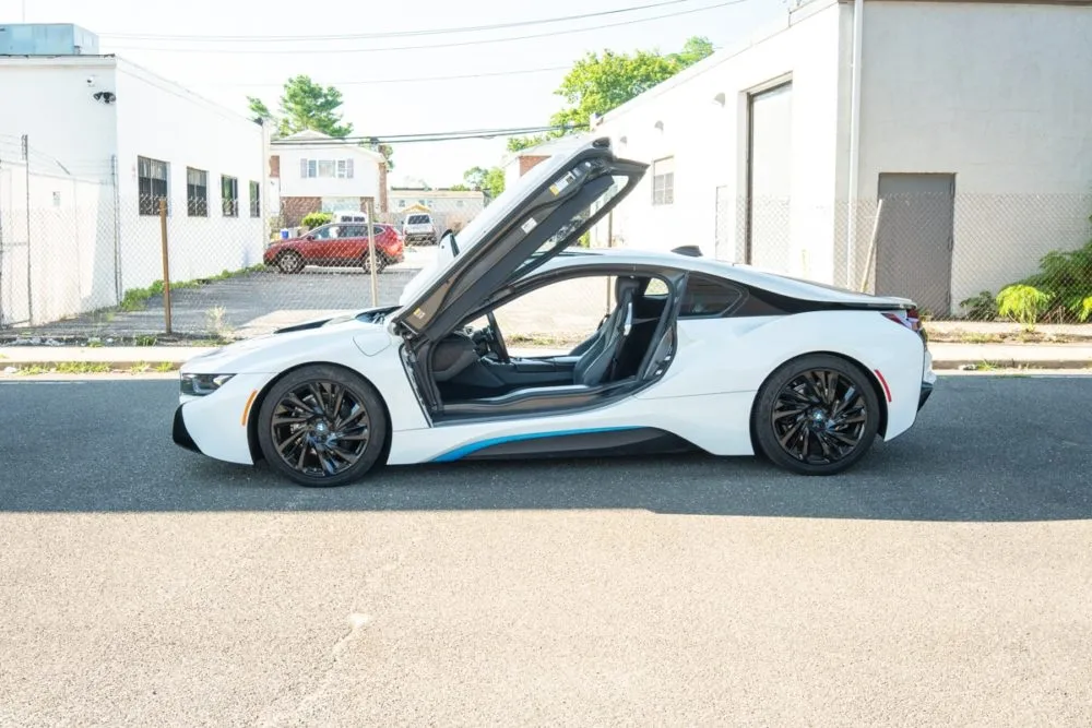 Side profile of a white BMW i8 with doors raised, showcasing its futuristic design and aerodynamic silhouette in bright daylight.