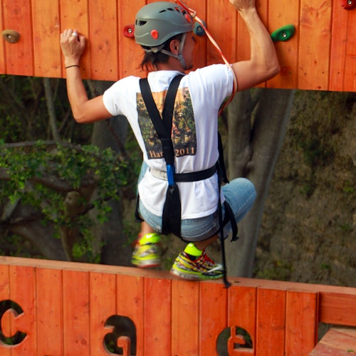 一个戴头盔和安全带的人正在攀爬木制障碍墙,穿着白色衬衫和霓虹色运动鞋。