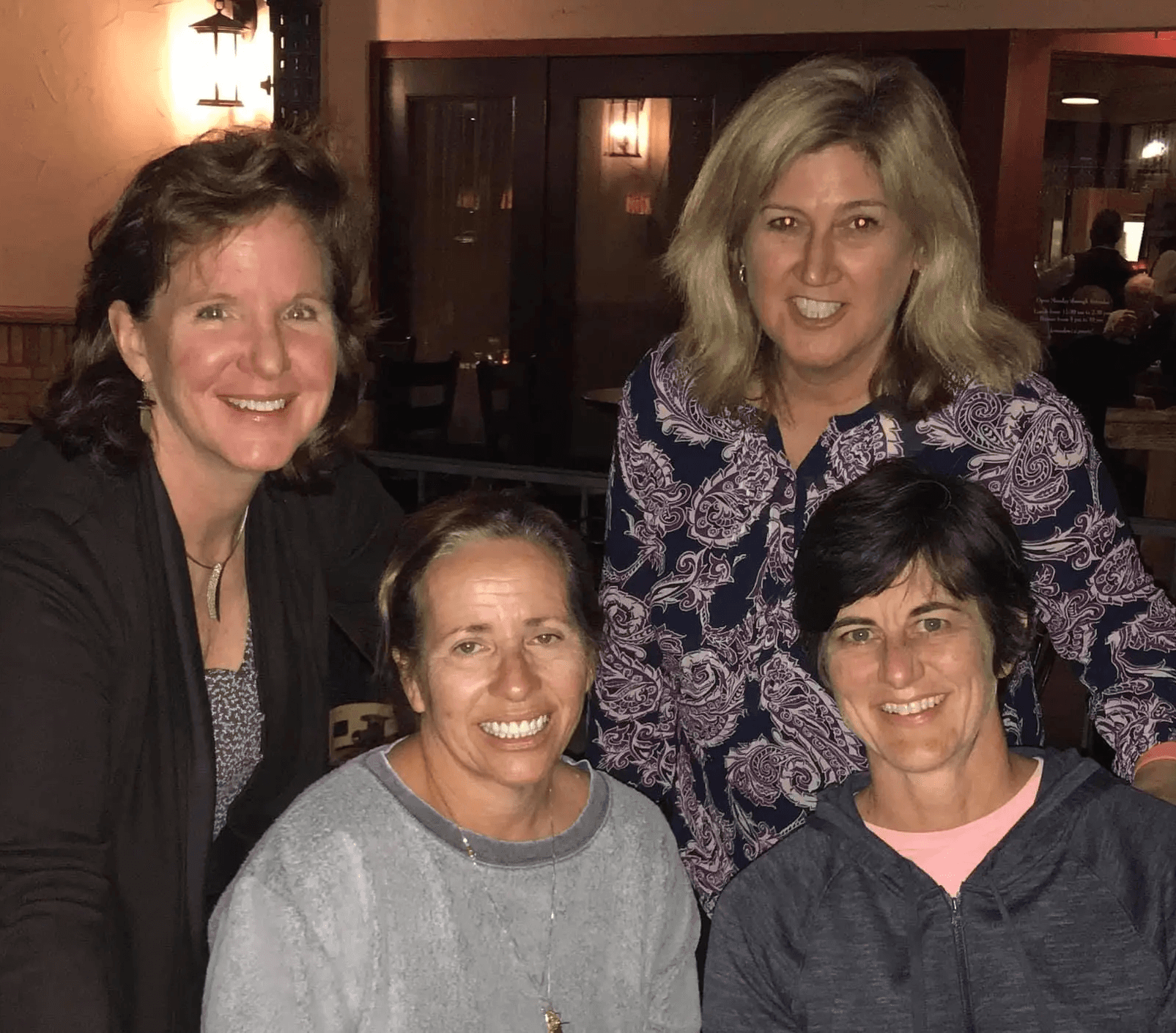 Four women are smiling and posing together in a warmly lit indoor setting, with wooden accents and soft lighting creating a cozy atmosphere.