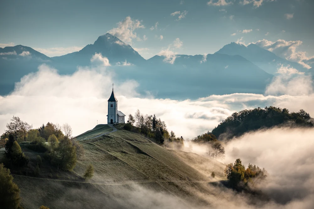 The hilltop Jamnik church in Slovenia at sunrise, framed by fog and a view of the distant Alps.
