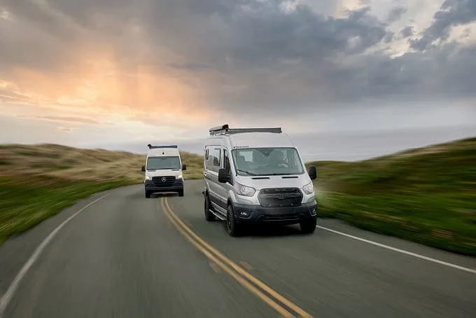An ambulance drives on a winding road under a cloudy sky at sunset, with green fields on either side.