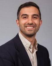 Smiling man in a navy blazer and checkered button-down shirt. Short dark hair and a friendly expression.