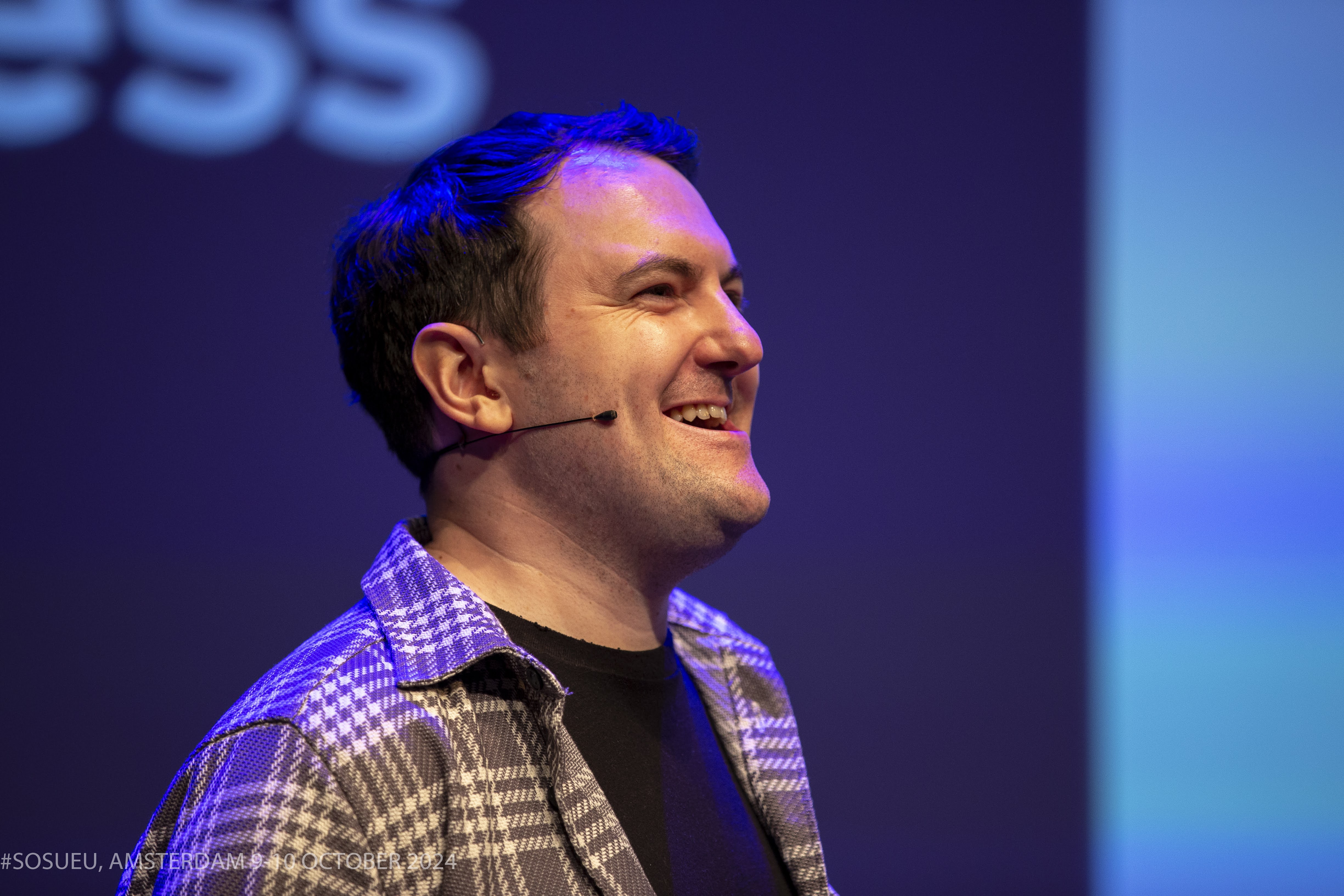 Matt Mercer smiles on stage, wearing a checkered shirt, with colorful lights in the background.