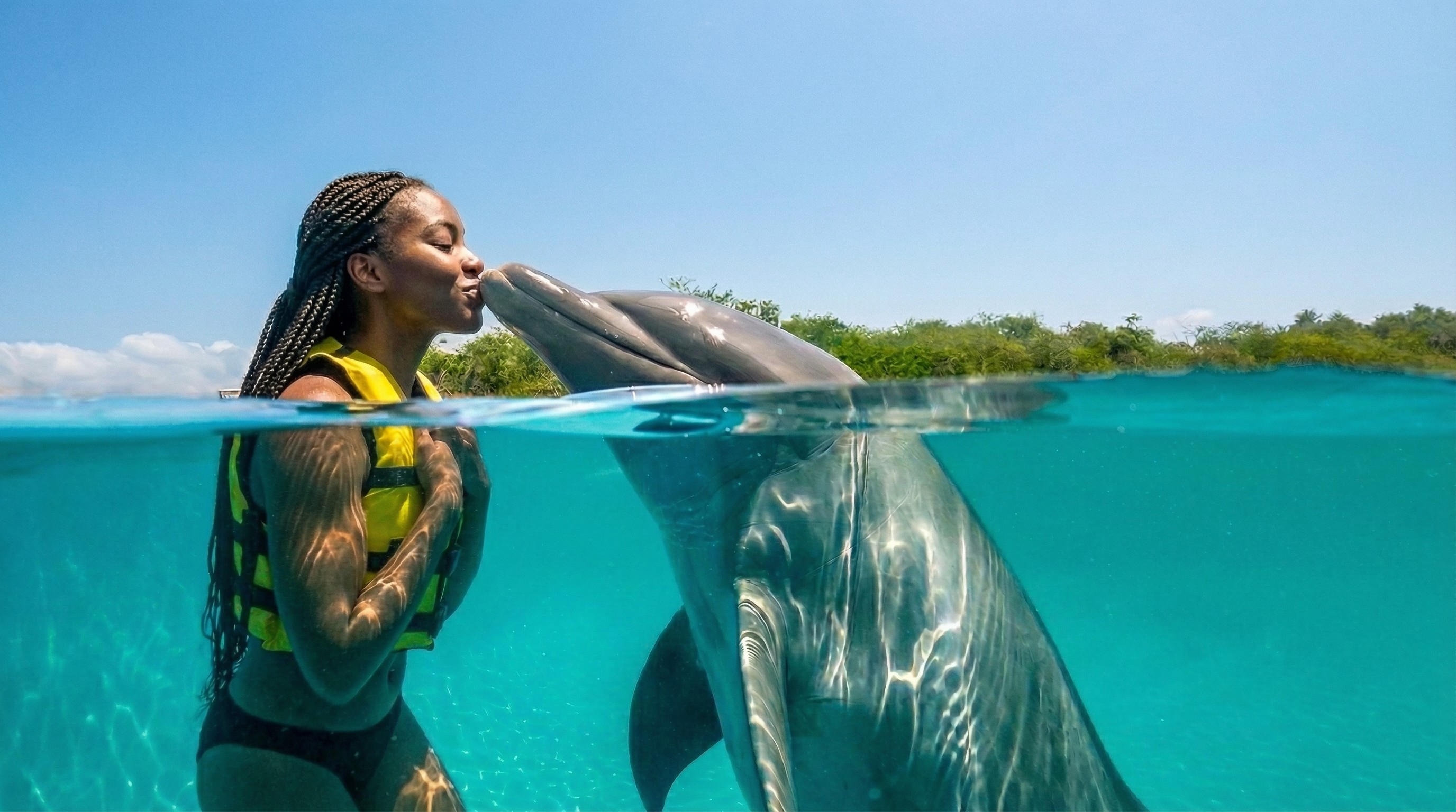 Woman in life vest kissing a dolphin in clear blue water.