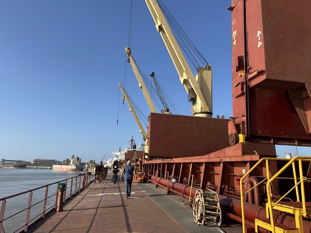 Crew walking along a bulk carrier deck with cranes and open hatch covers alongside a terminal — on-the-ground port operations and cargo-ready maritime execution.